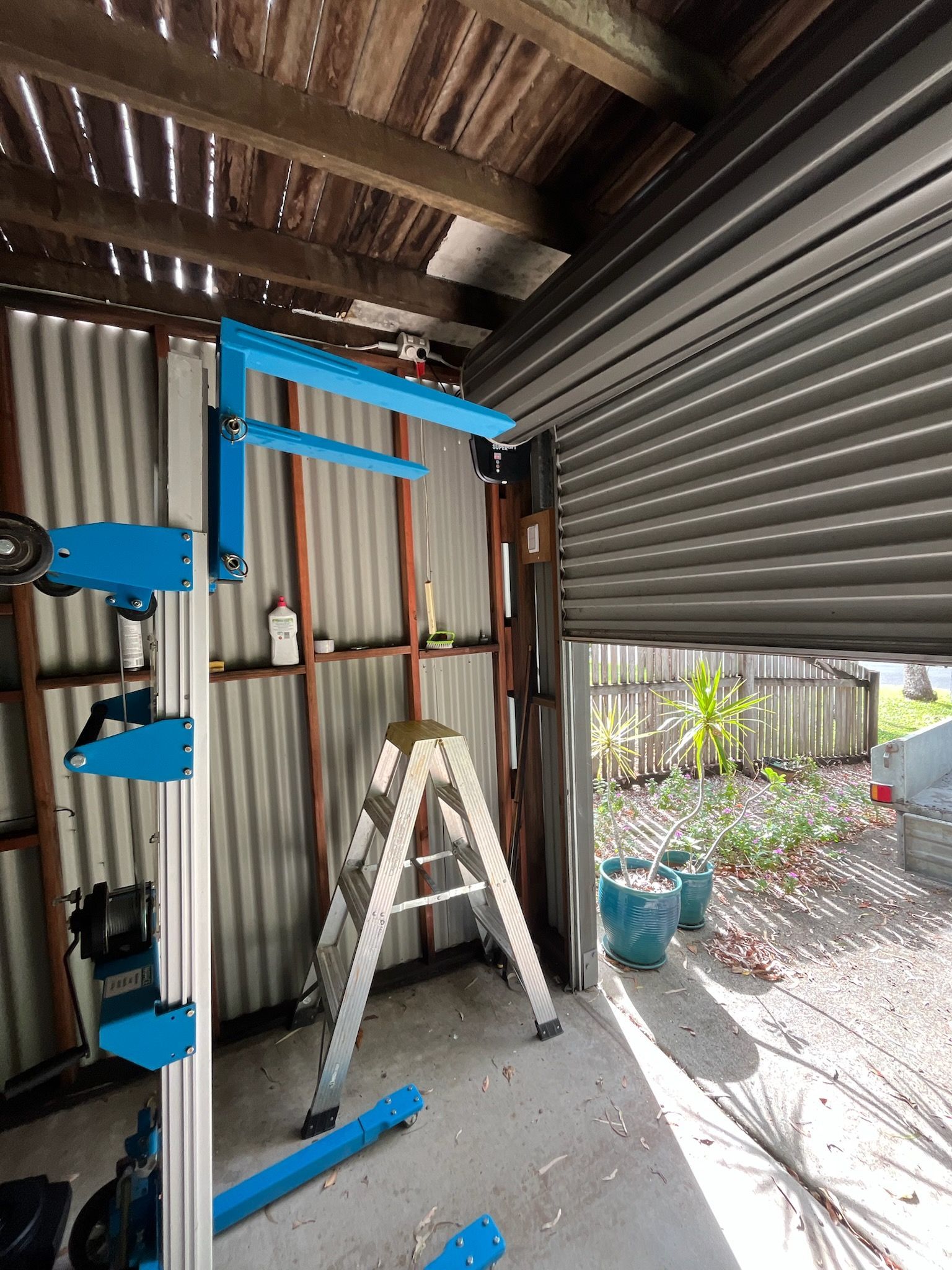 Blue lifting equipment and a white ladder inside a corrugated metal shed, with an open doorway outside — Rolling With Netherpoel in Proserpine, QLD