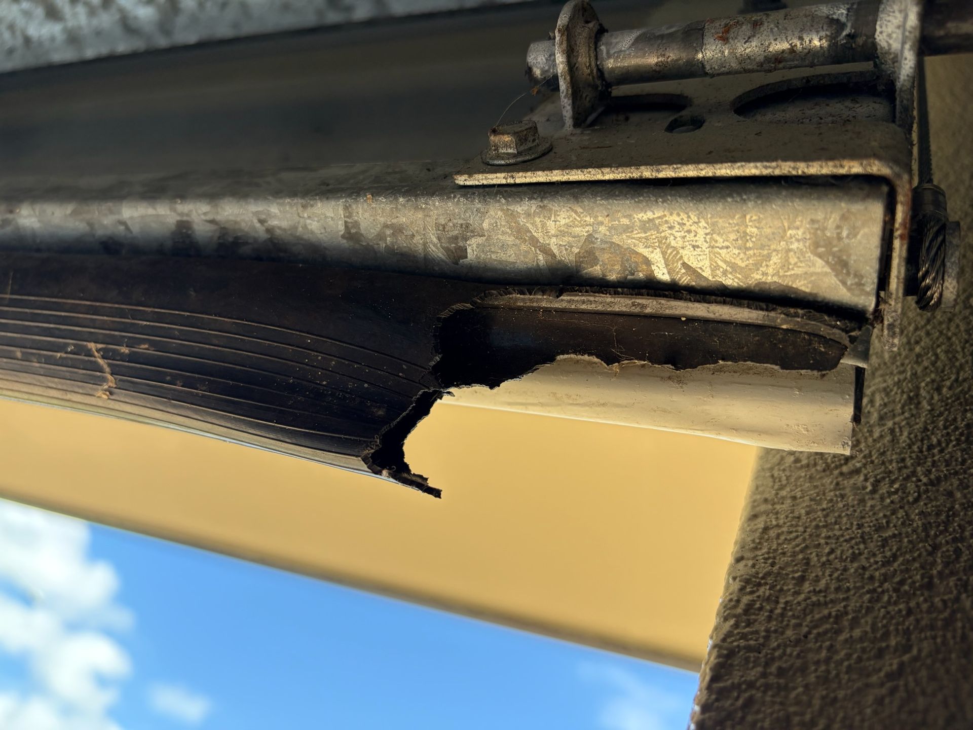 Close-up of a torn black awning edge with a metal support bracket against a beige wall and blue sky — Rolling With Netherpoel in Proserpine, QLD