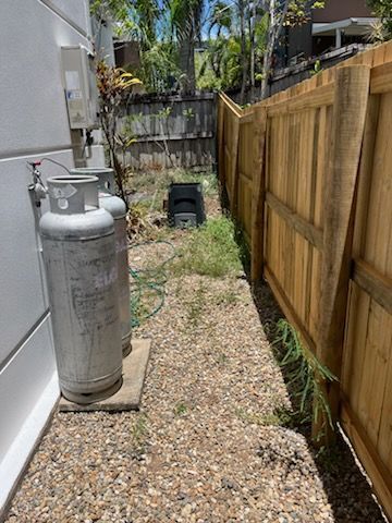 Narrow gravel side yard with a silver propane tank, wooden fence, and small black bin near the back — Rolling With Netherpoel in Cannonvale, QLD