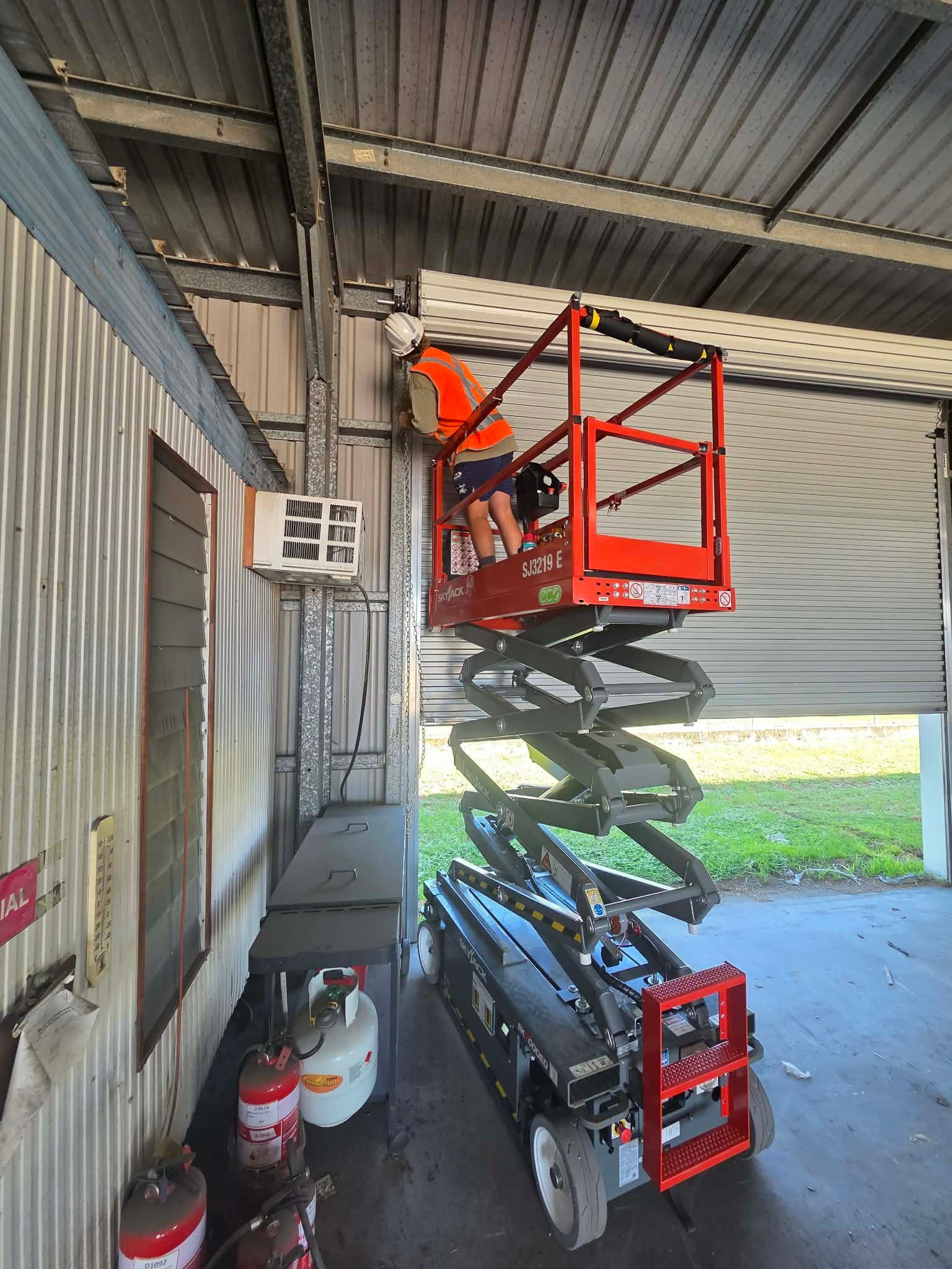 Worker in an orange vest on a scissor lift inside a covered loading area, adjusting overhead metal panels — Rolling With Netherpoel in Bowen, QLD