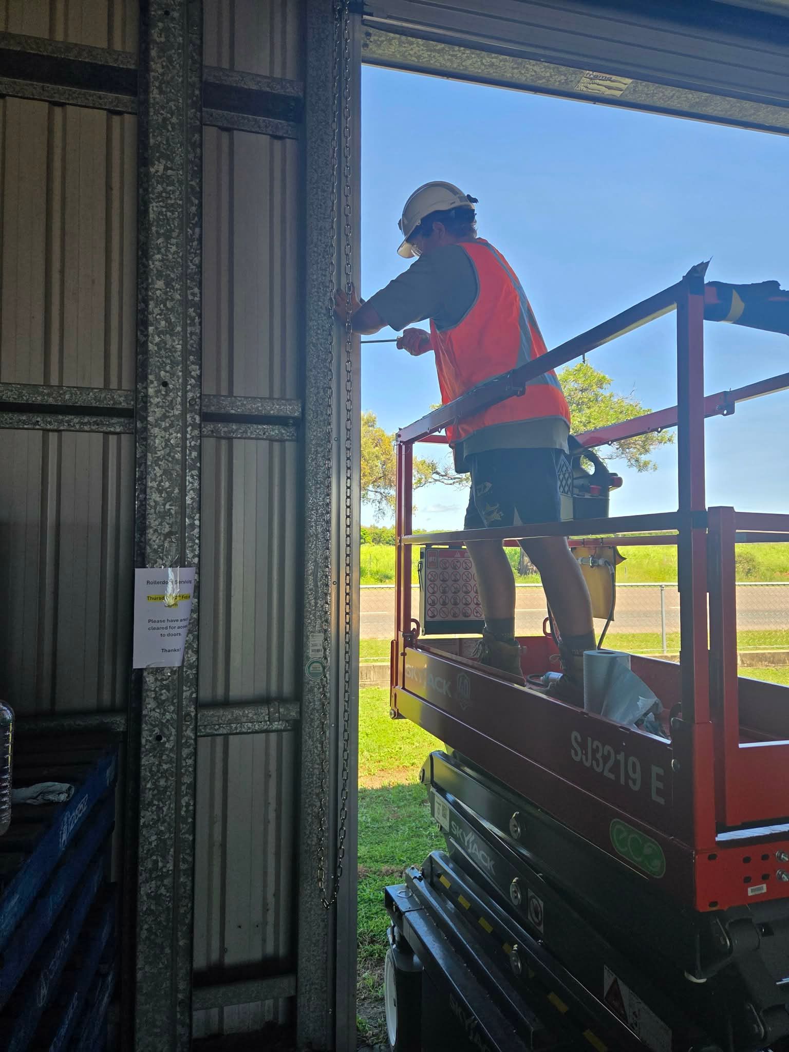 Worker in orange vest and hard hat standing on a scissor lift, reaching into a metal building doorway — Rolling With Netherpoel in Bowen, QLD
