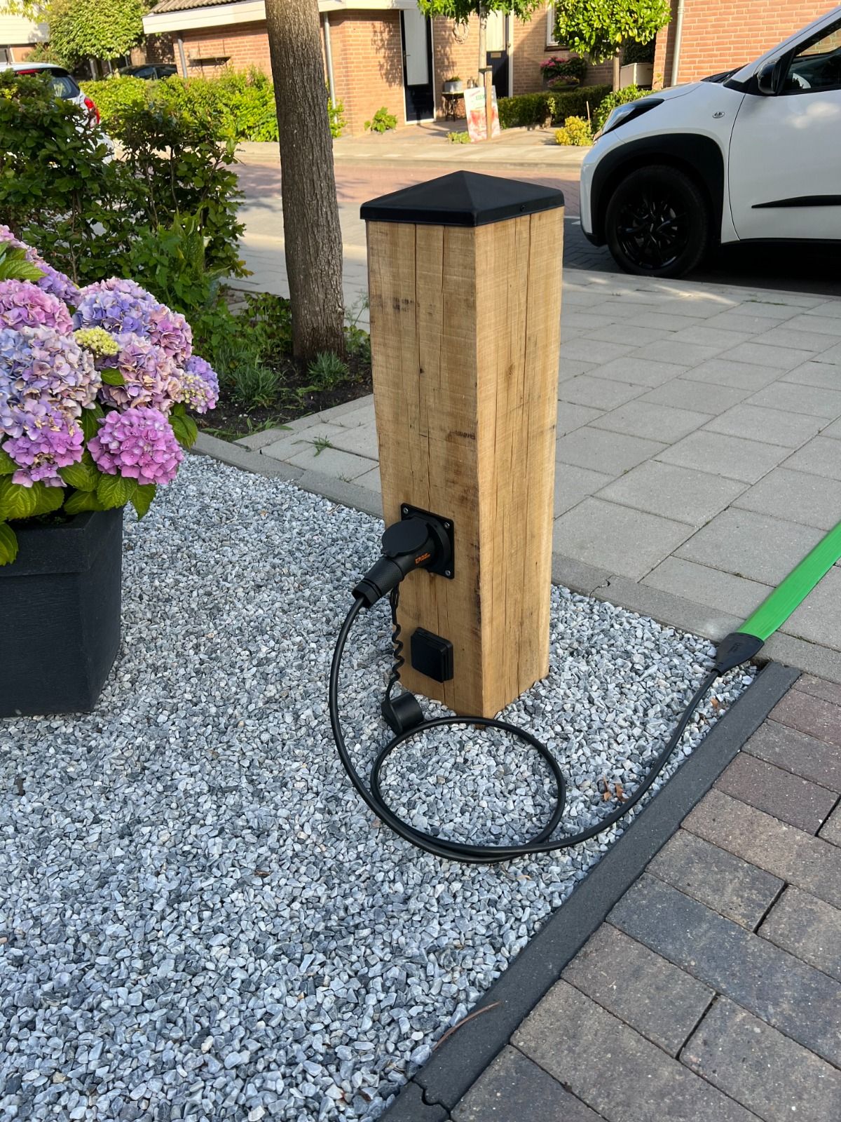 Houten laadpaal voor elektrische auto's in een grindbed naast een hortensiastruik, met een auto op de achtergrond.