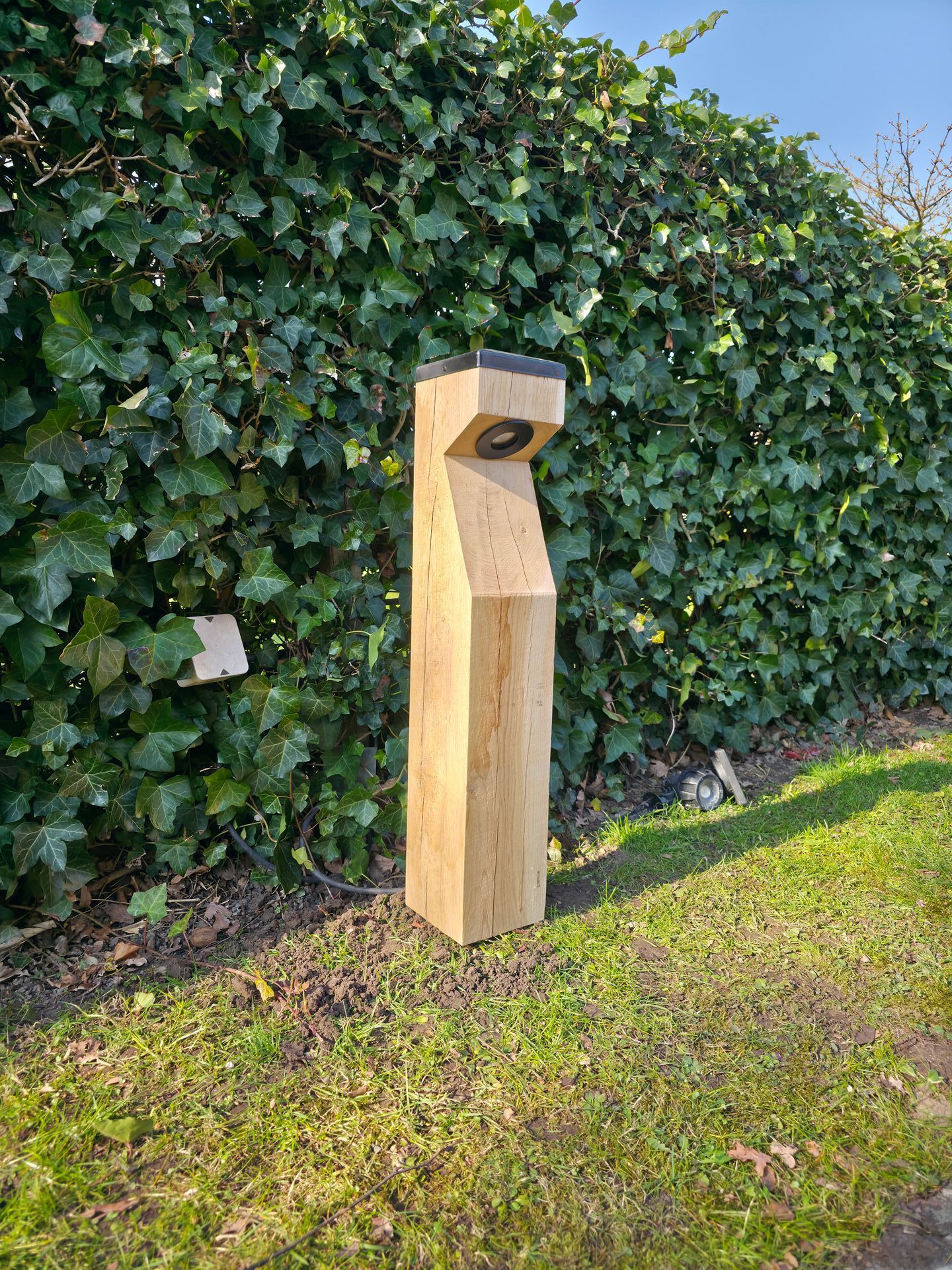 Houten buitenlamp met een schuin ontwerp, een zwarte bovenkant en een ronde lamp, geplaatst in een grasveld met groen.
