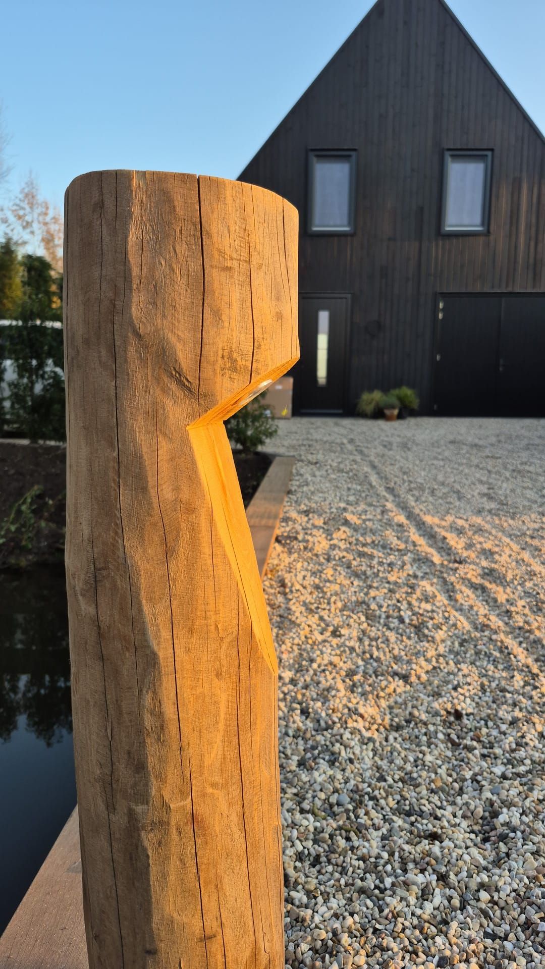 Houten paal met hoekige uitsparing, voor een modern zwart huis op een grindpad.