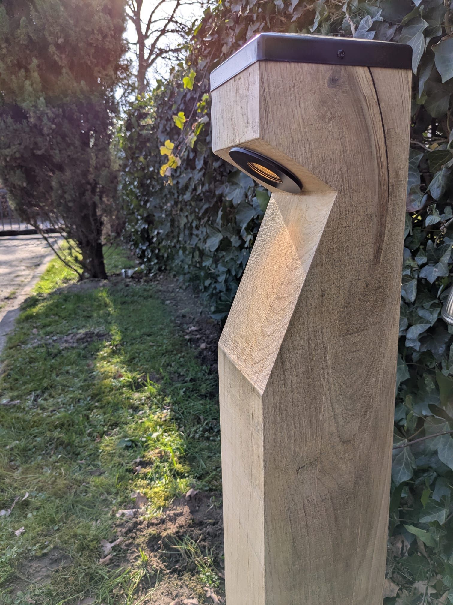 Houten buitenlamp met een schuin ontwerp, een zwarte bovenkant en een ronde lamp, geplaatst in een grasveld met groen.
