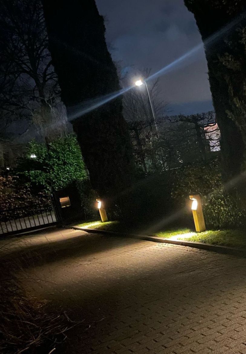 Nachtscène van een pad verlicht door gele grondverlichting en een straatlantaarn. Aan de zijkanten staan ​​bomen.