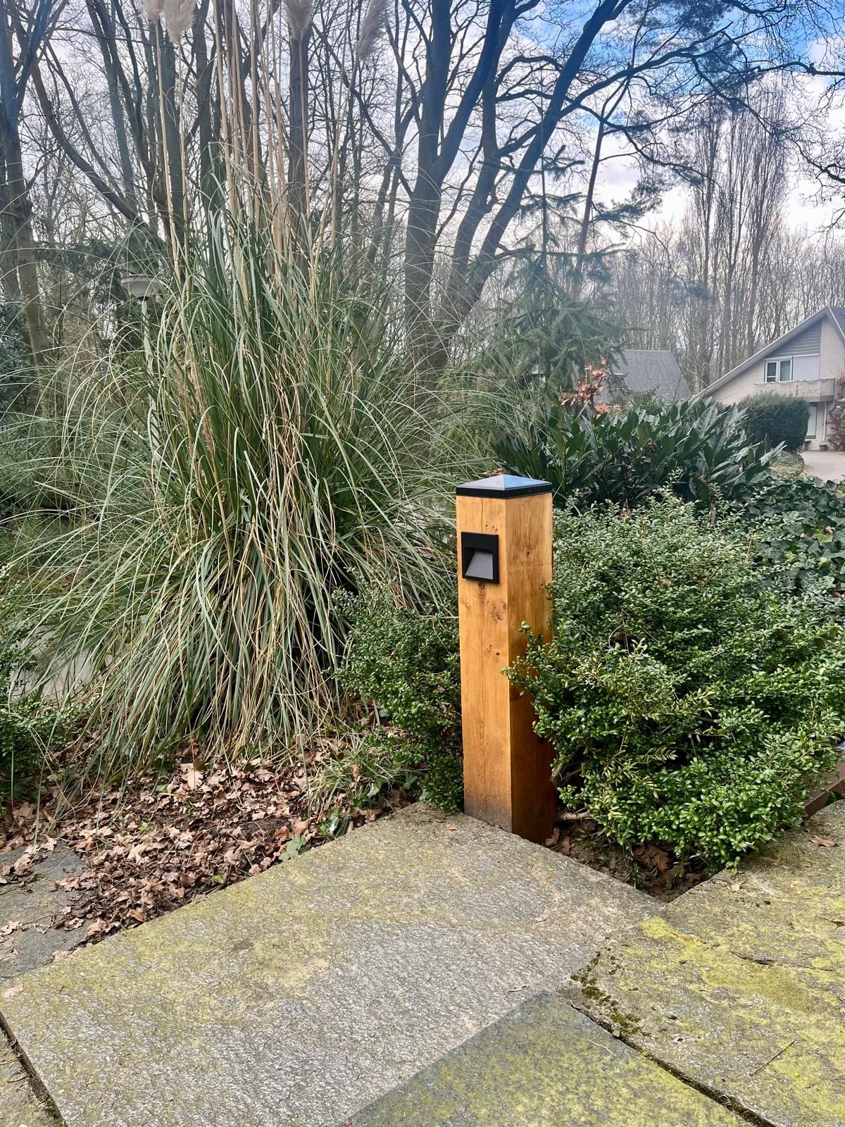 Een houten paal, mogelijk een lamp, in een bloembed met weelderig groen, een betonnen pad en een huis op de achtergrond.