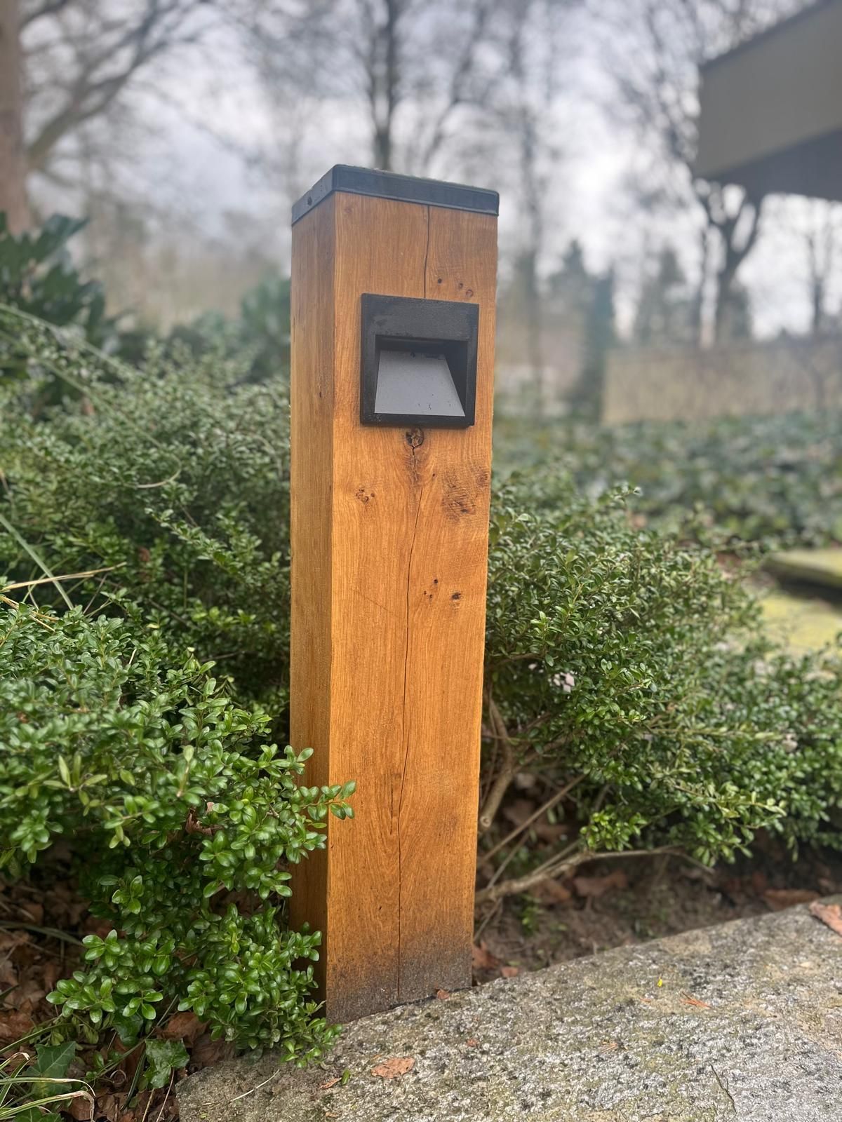 Houten paal met een zwarte vierkante lamp in een tuin.