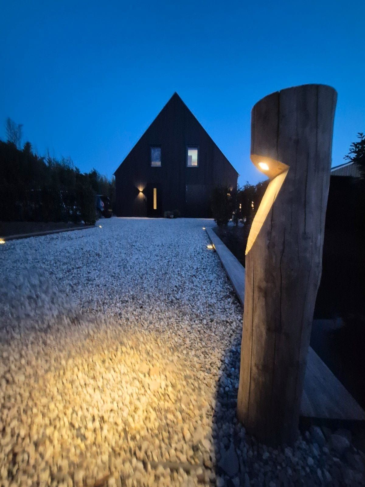 Houten paal met hoekige uitsparing, voor een modern zwart huis op een grindpad.