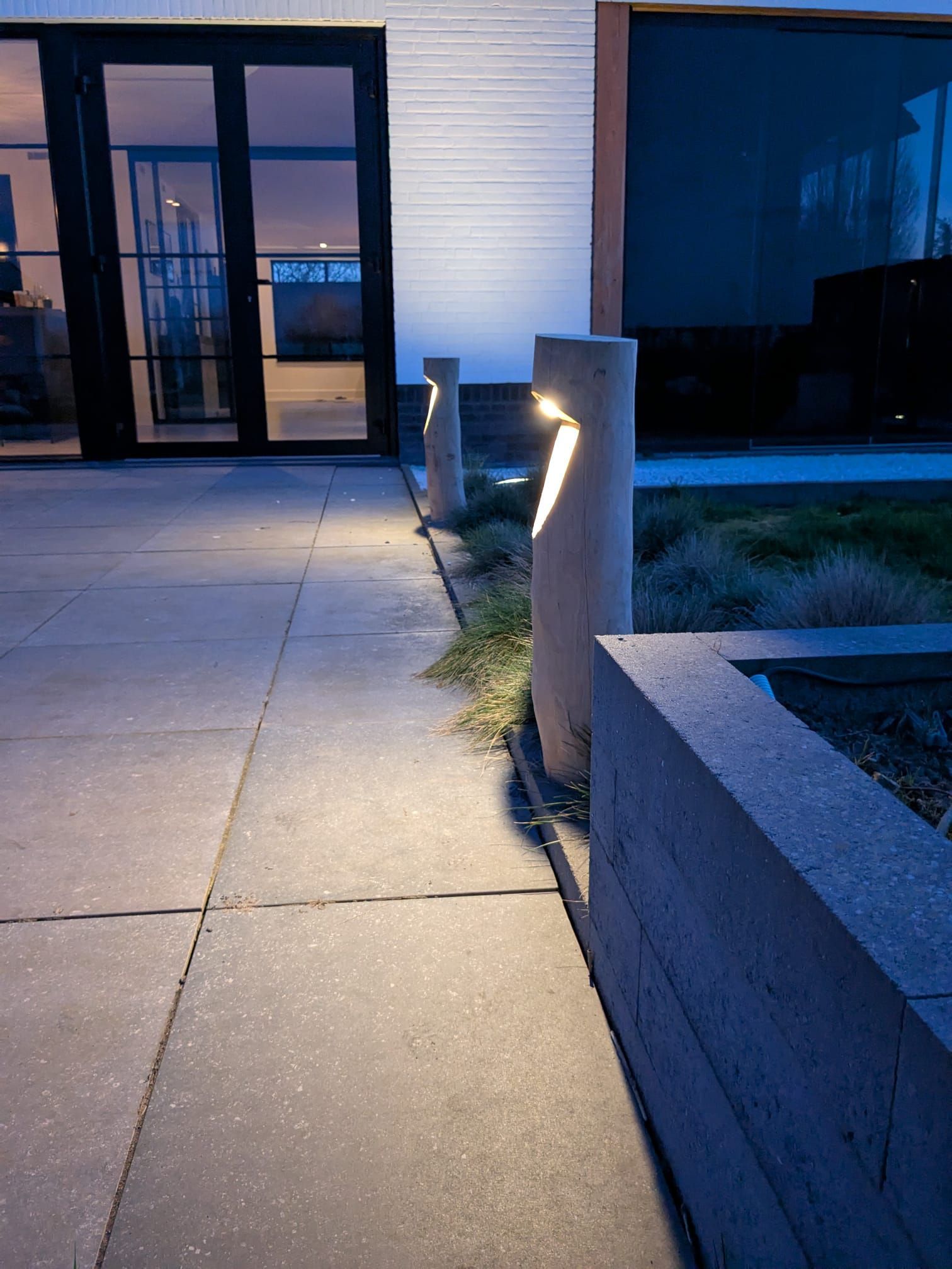 Een betonnen pad, verlicht door moderne pilaarlampen, leidt naar glazen deuren in een gebouw bij schemering.