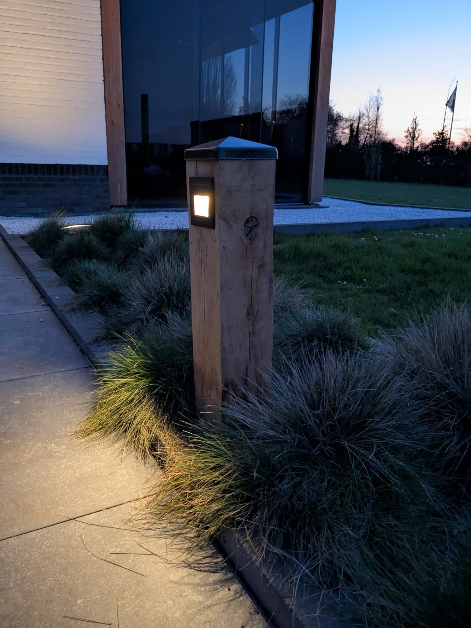 Houten paal met een vierkante lamp, die het voetpad en de planten voor een gebouw verlicht.