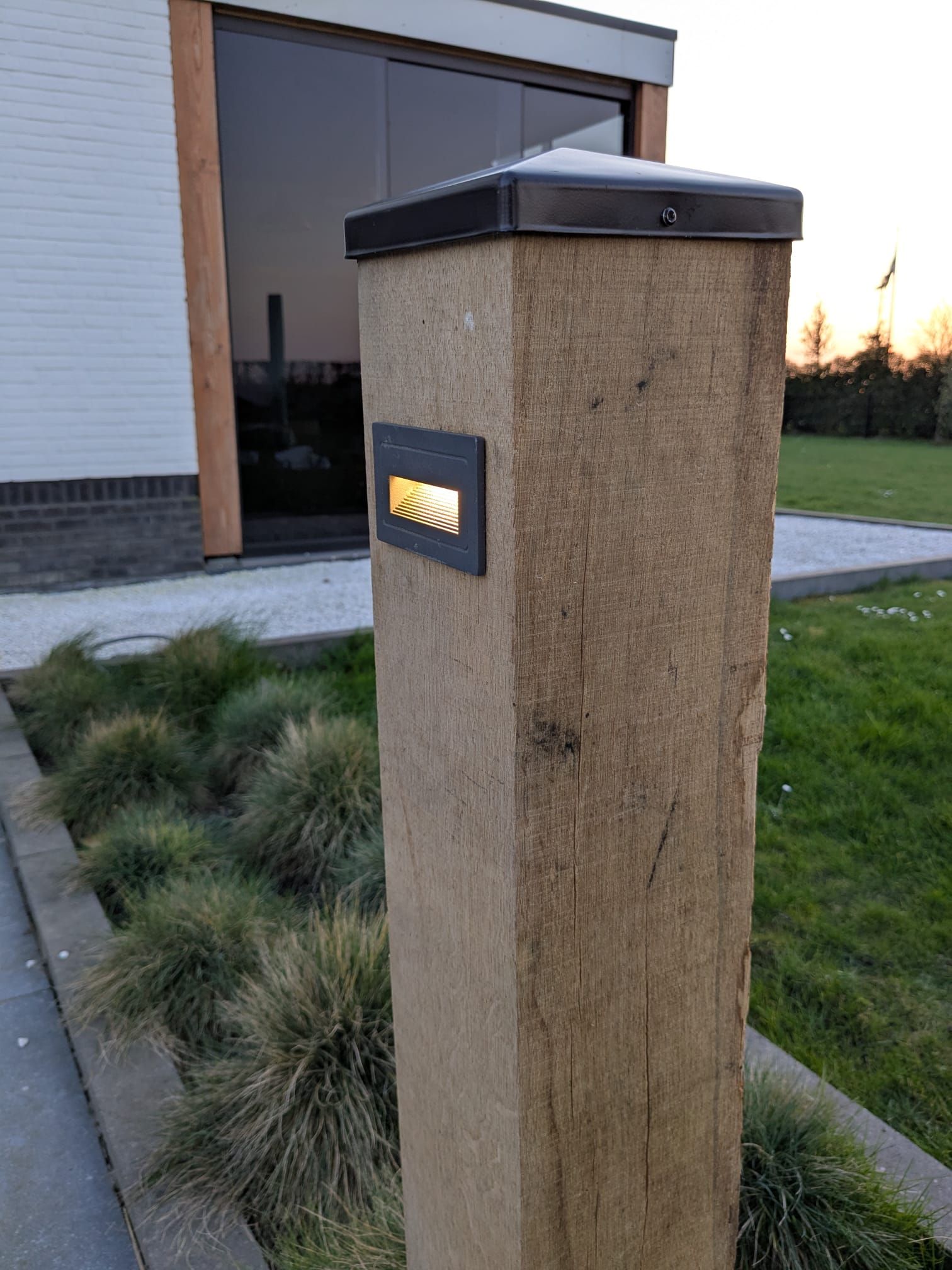 Houten paal met ingebouwde verlichting in een bloembed naast een wandelpad en een modern gebouw.