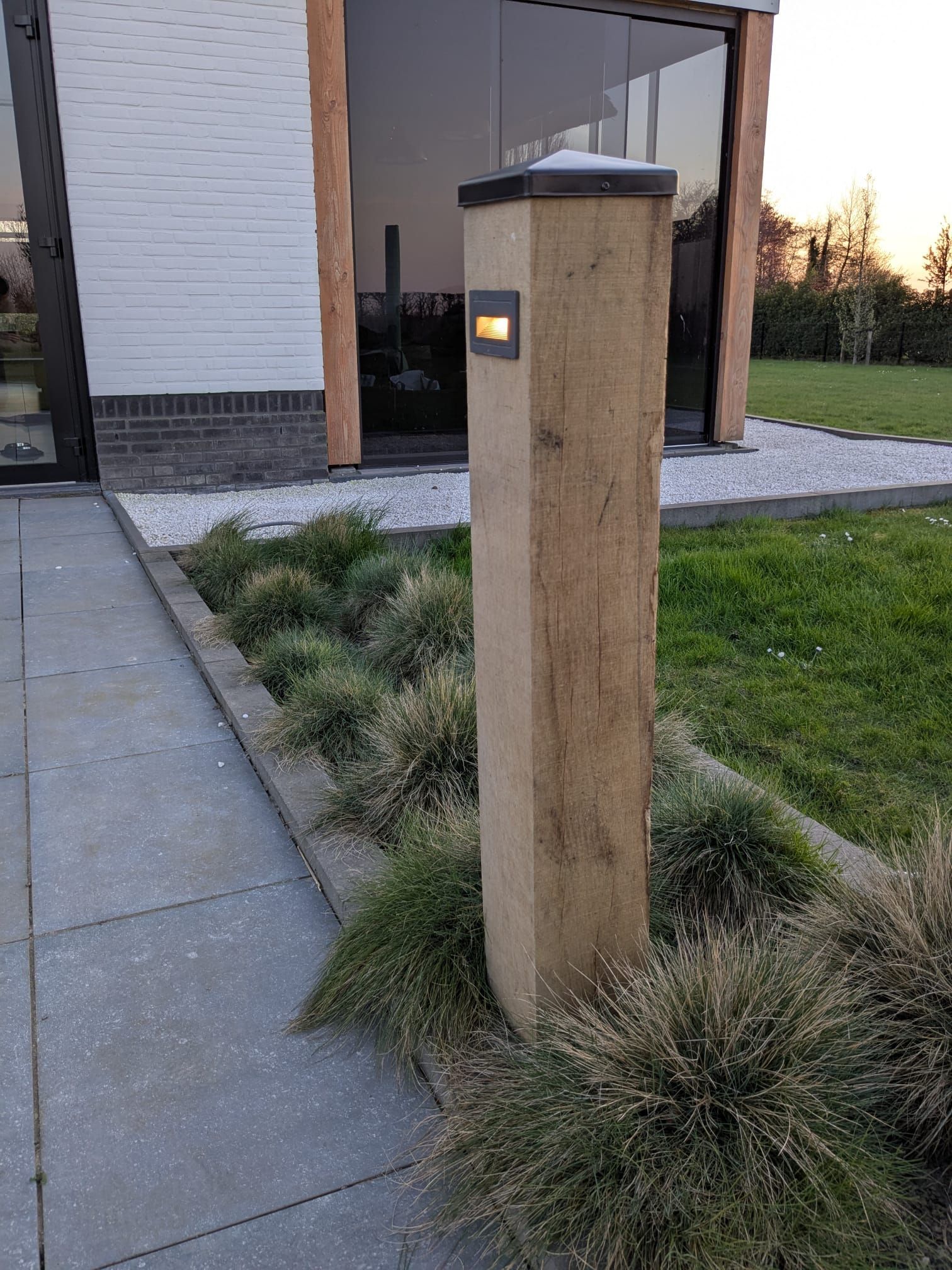 Houten paal met ingebouwde verlichting in een bloembed naast een wandelpad en een modern gebouw.