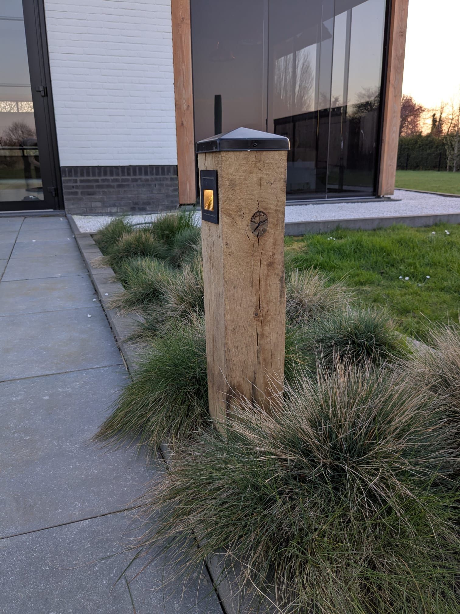 Houten paal met ingebouwde brievenbus, omgeven door siergras en grenzend aan een betonnen pad.