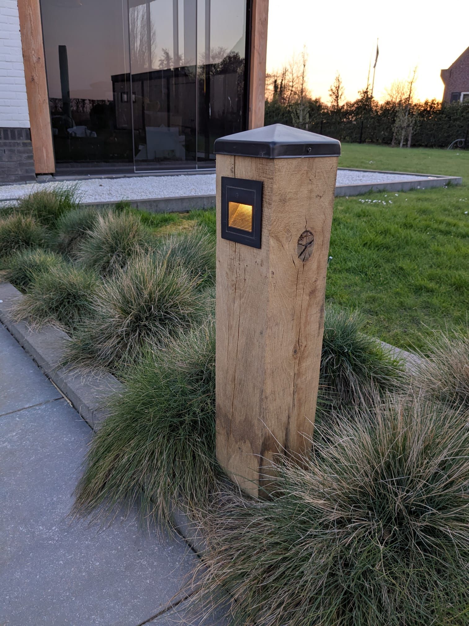 Houten paal met ingebouwde brievenbus, omgeven door siergras en grenzend aan een betonnen pad.