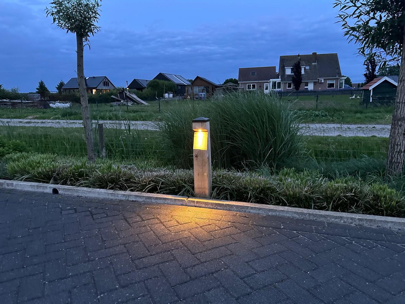 Een verlichte lantaarnpaal op een bakstenen pad, met huizen en gras op de achtergrond onder een schemerige hemel.