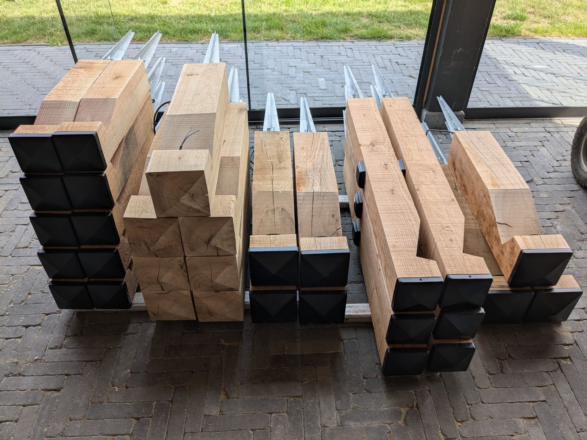 Stapels houten palen met zwarte piramidevormige doppen, op een grijs terras. Metalen steunen bevestigd.