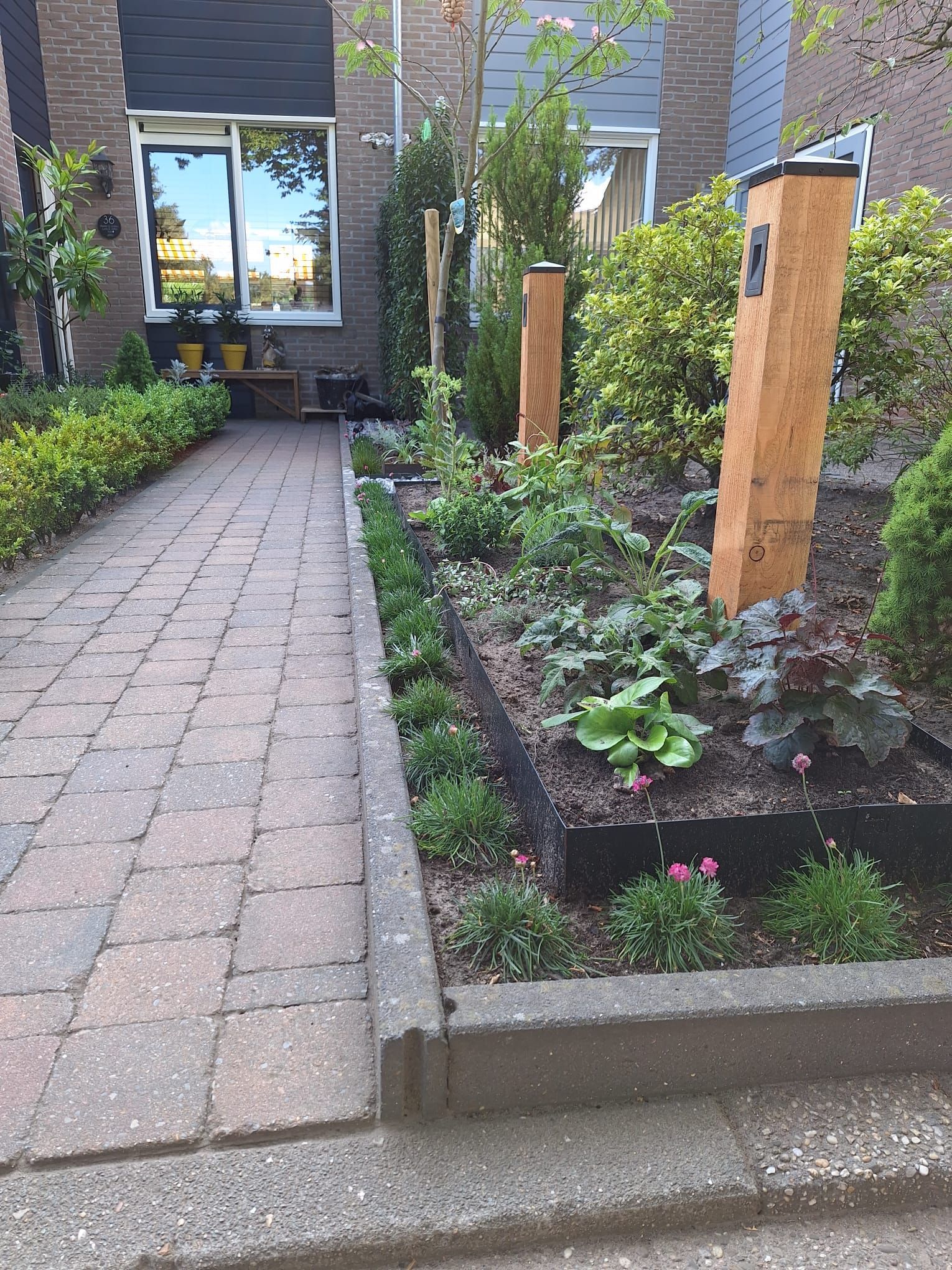 Een bakstenen pad loopt langs een verhoogd bloembed met planten, houten palen en een huisje.