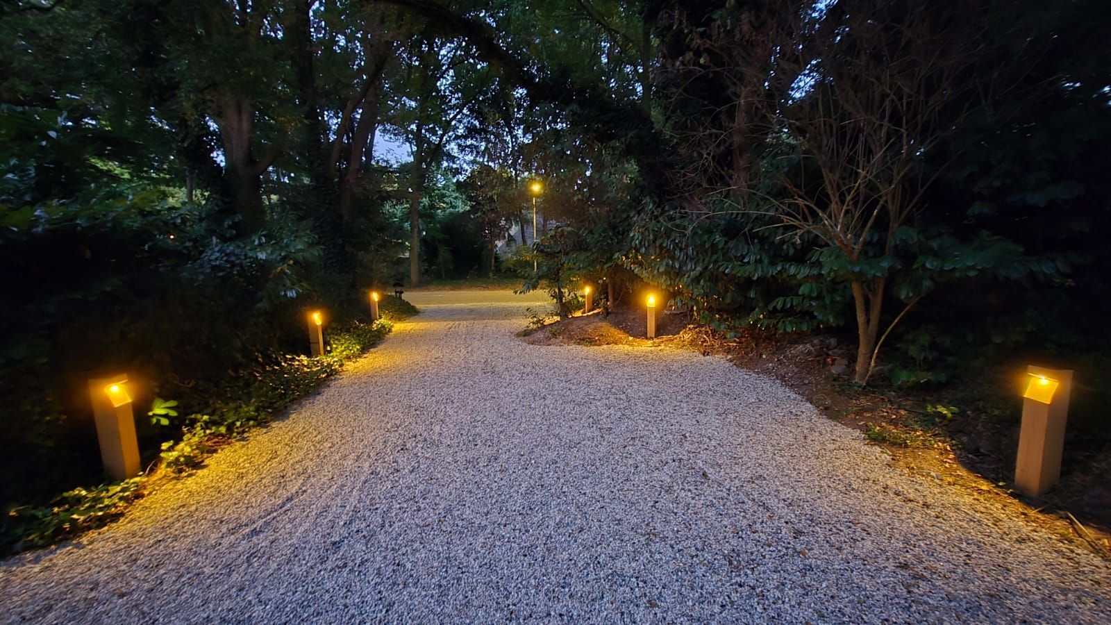 Een grindpad, verlicht door gouden lampen en omzoomd met groen, in de schemering.
