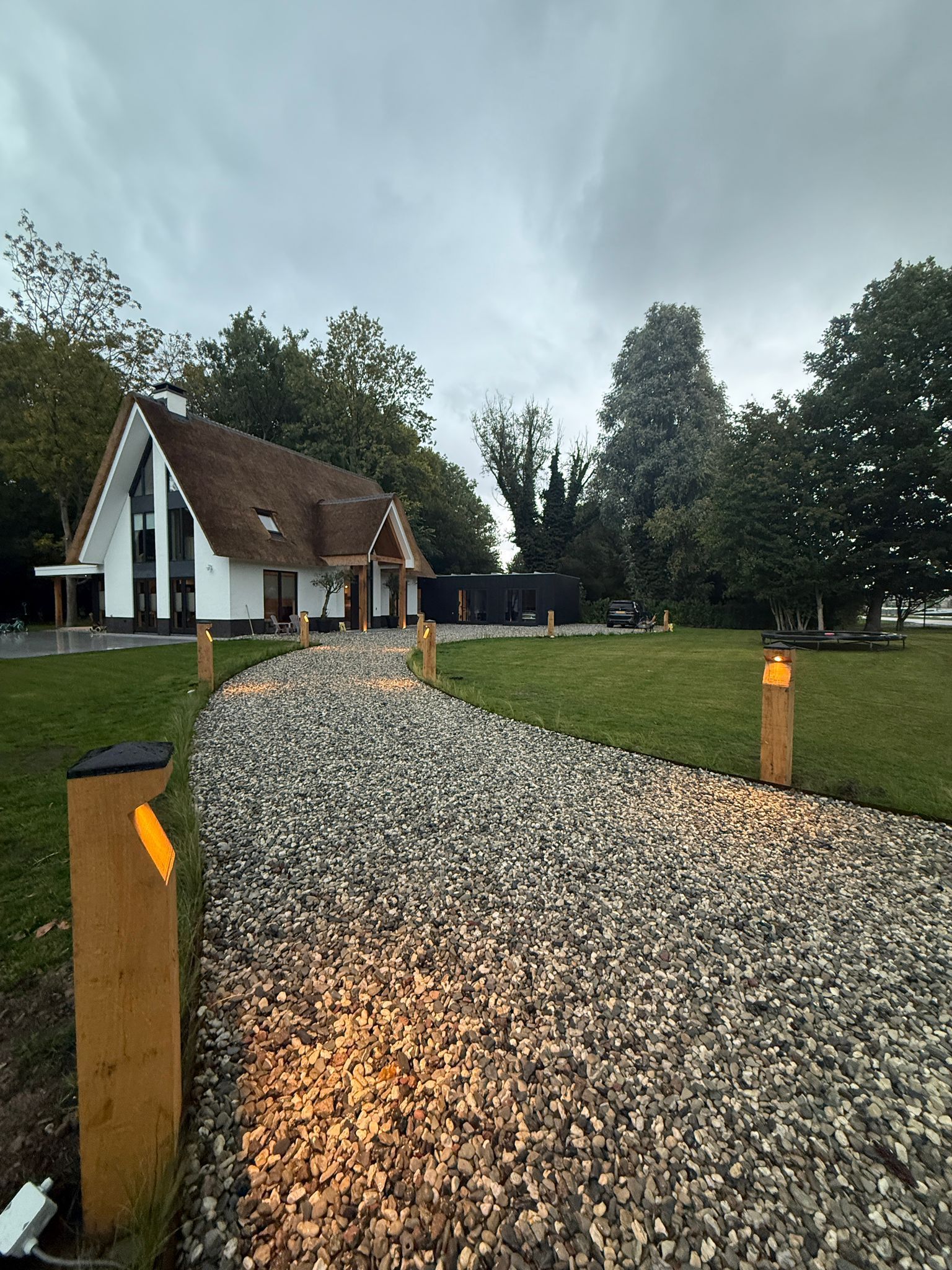 Een grindpad, verlicht met houten lantaarns, leidt naar een wit huis met een bruin dak onder een bewolkte hemel.
