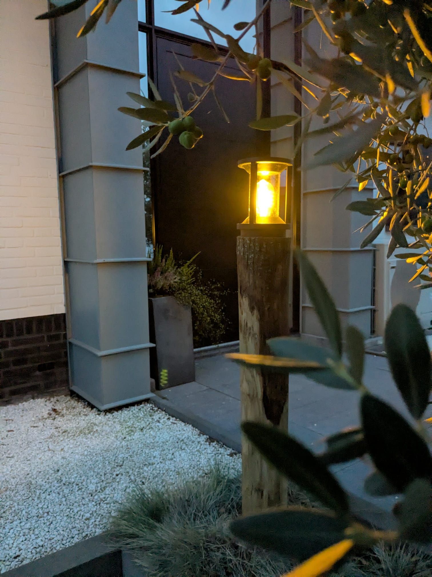 Een verlichte buitenlantaarnpaal met een felle lamp, naast een wit gebouw onder een boom.