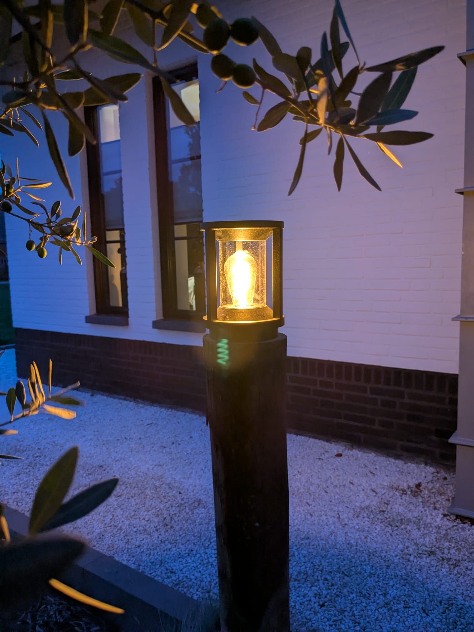 Een verlichte buitenlantaarnpaal met een felle lamp, naast een wit gebouw onder een boom.