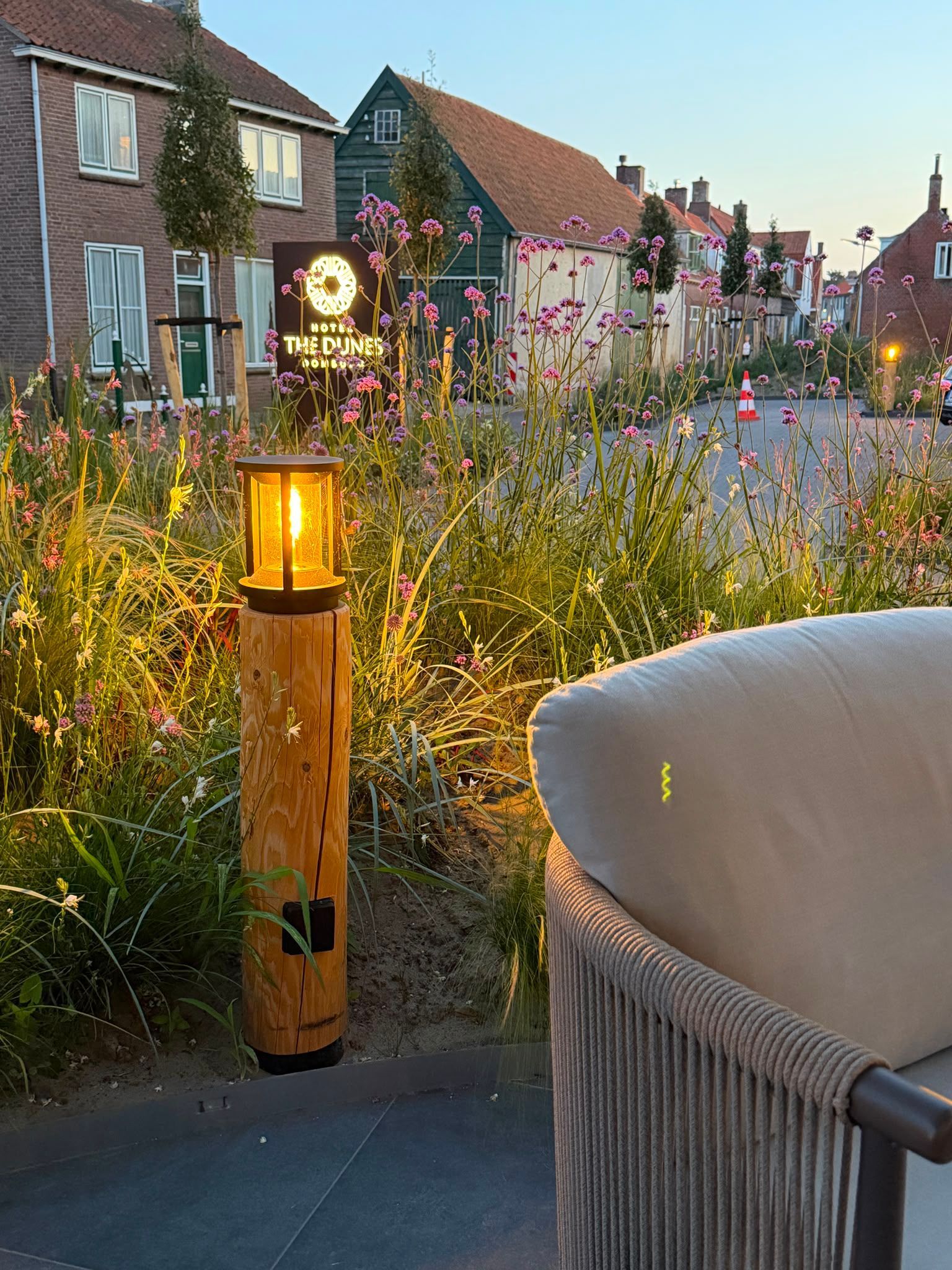 Een brandende lantaarn op een verweerde houten paal in een tuin, naast een olijfboom.