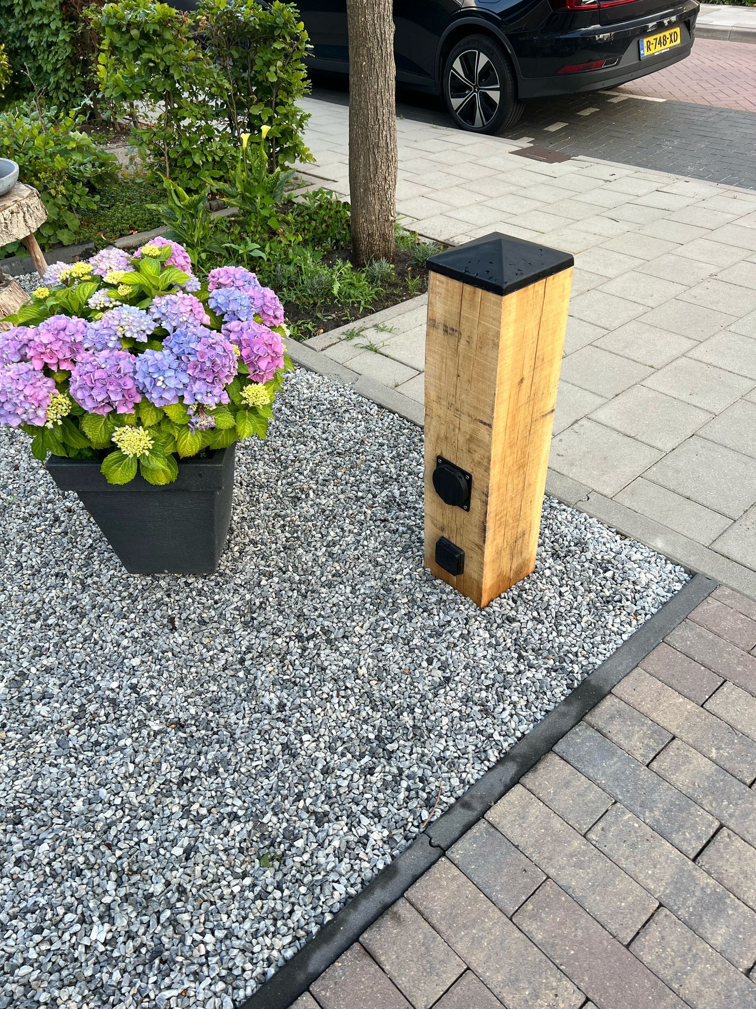 Een laadstation voor elektrische voertuigen van hout, naast een verharde oprit en een hortensia.