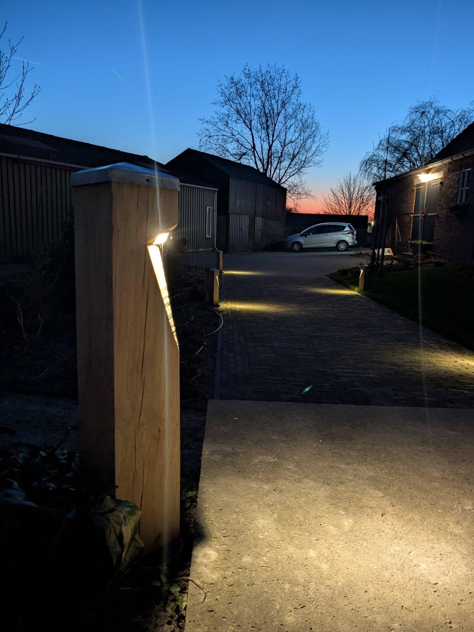 Verlicht pad met houten palen, in de schemering. Op de achtergrond zijn een auto en een schuur te zien.