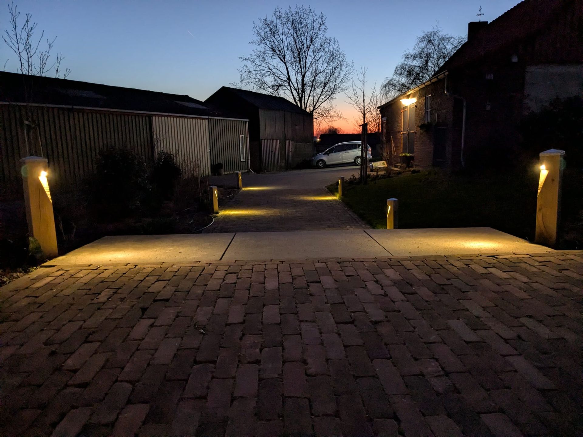 Een pad in de schemering, met bakstenen bestrating en verlichte pilaren, dat naar gebouwen leidt.