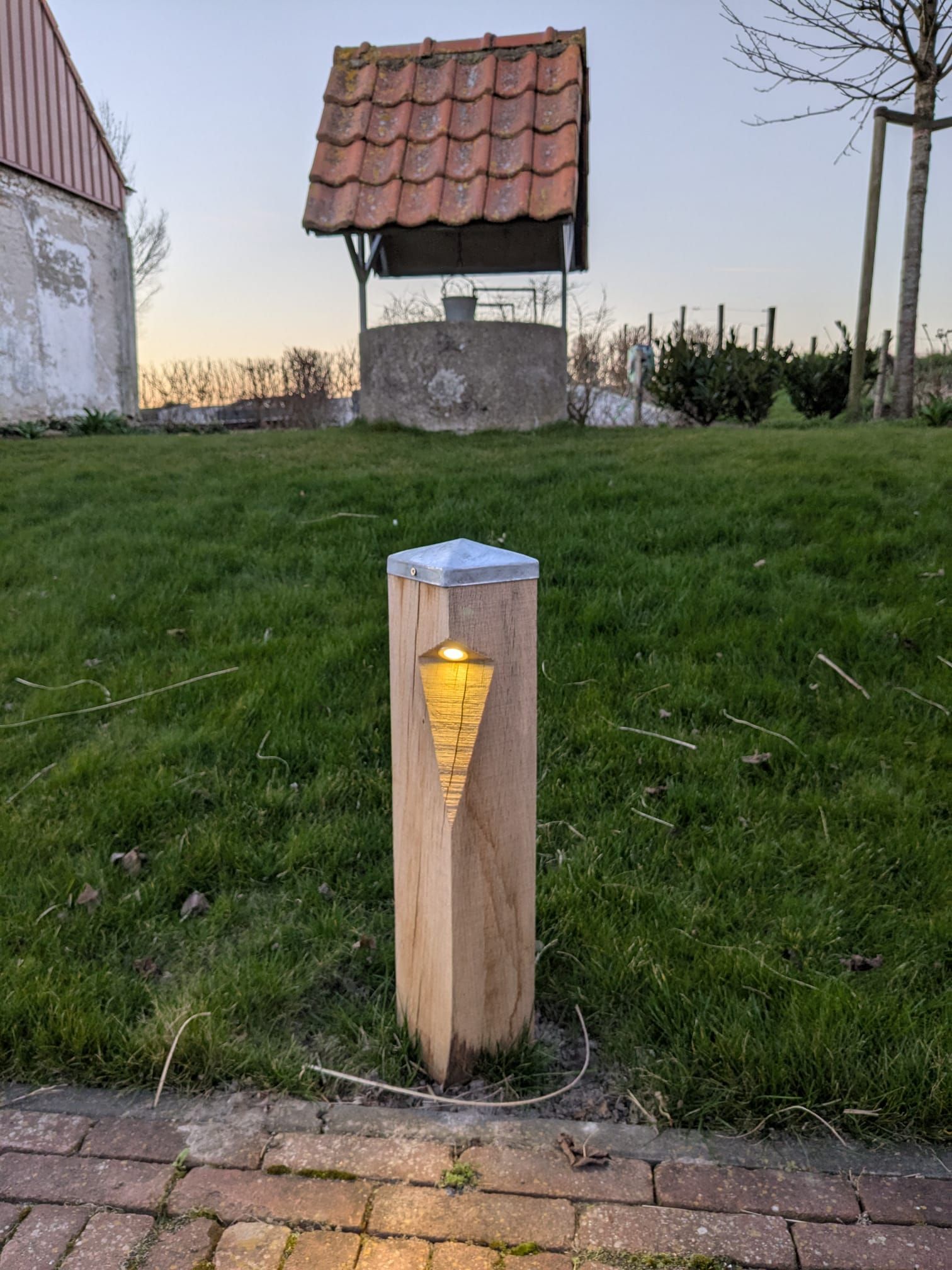 Houten paal met een gloeiend amberkleurig licht, voor een waterput op een grasheuvel, in de schemering.