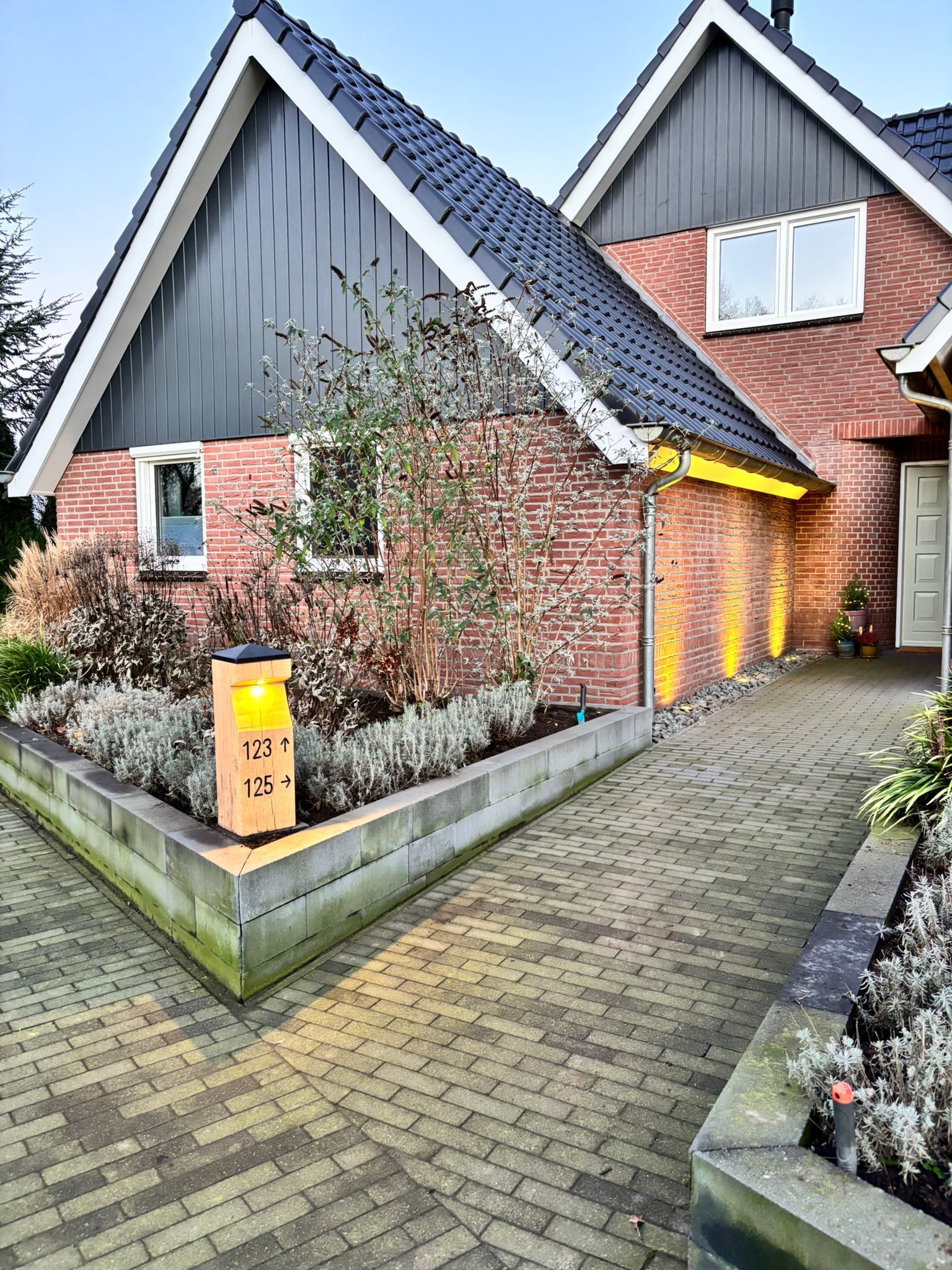 Houten adresbord met verlichte nummers 123 en 125, naast een bakstenen huis met een looppad.