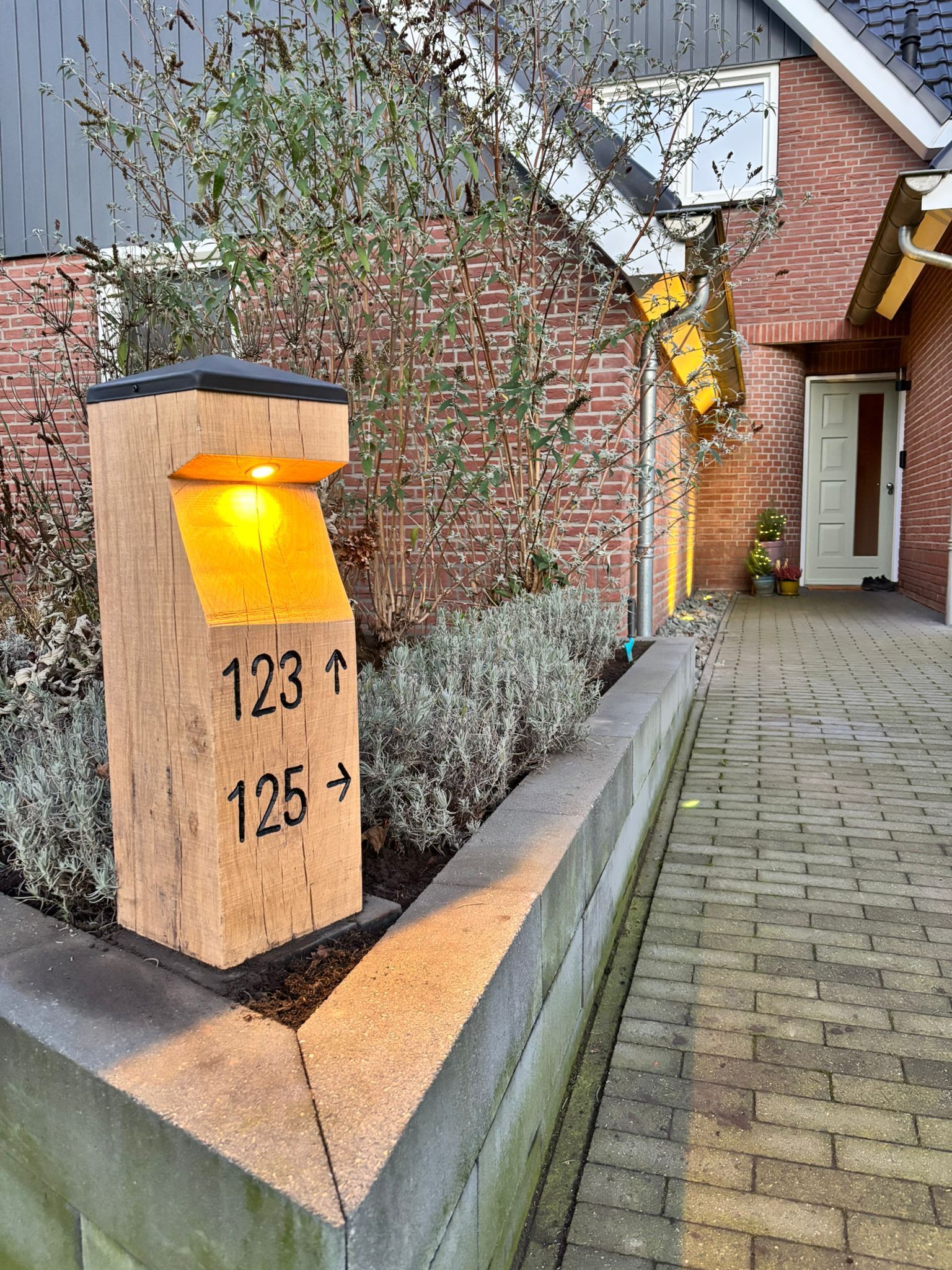 Houten paal met verlichte huisnummers 123 en 125, geplaatst in een tuin met een bakstenen huis op de achtergrond.