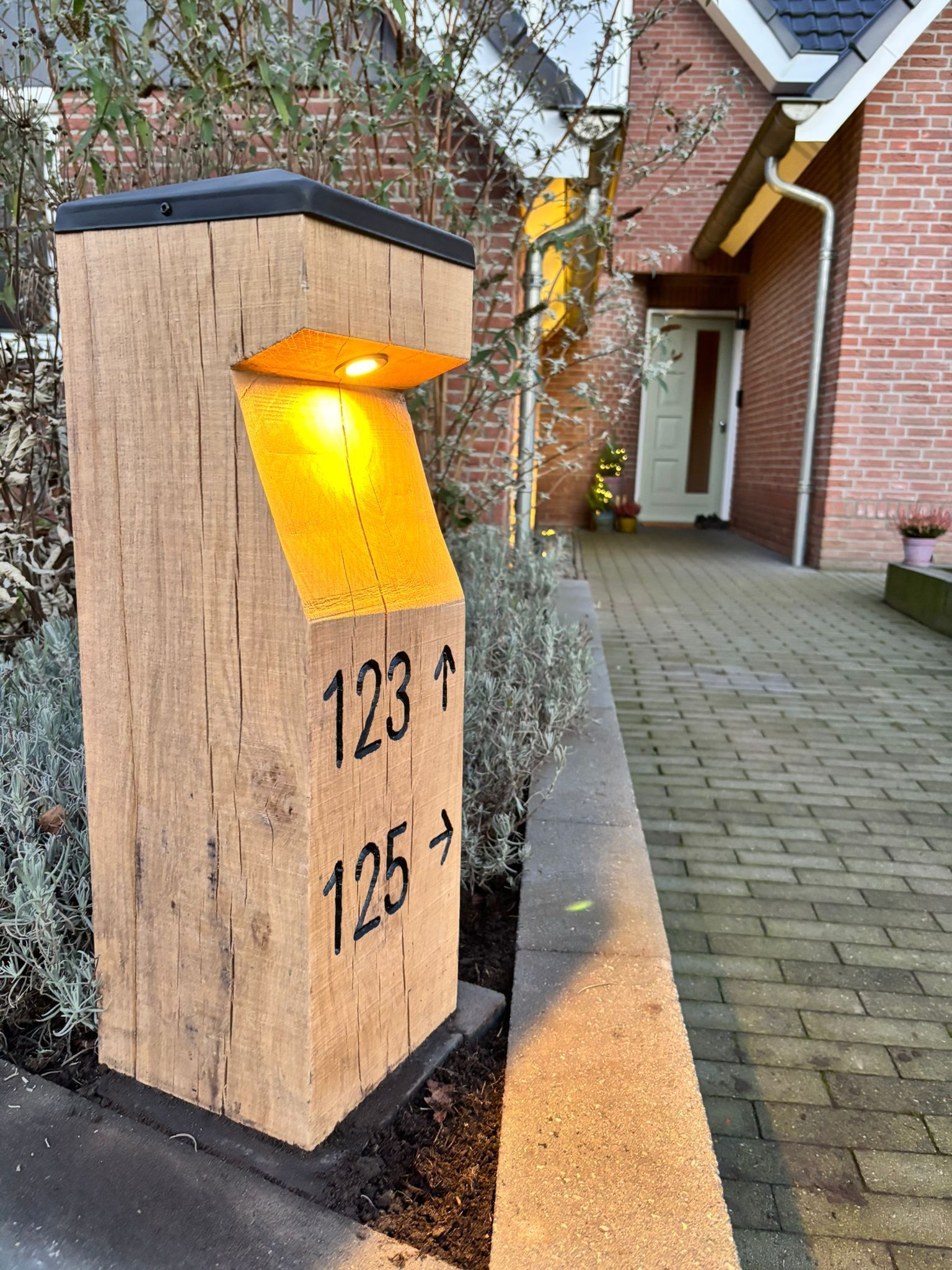 Houten adresbord met verlichte nummers 123 en 125, naast een bakstenen huis met een looppad.
