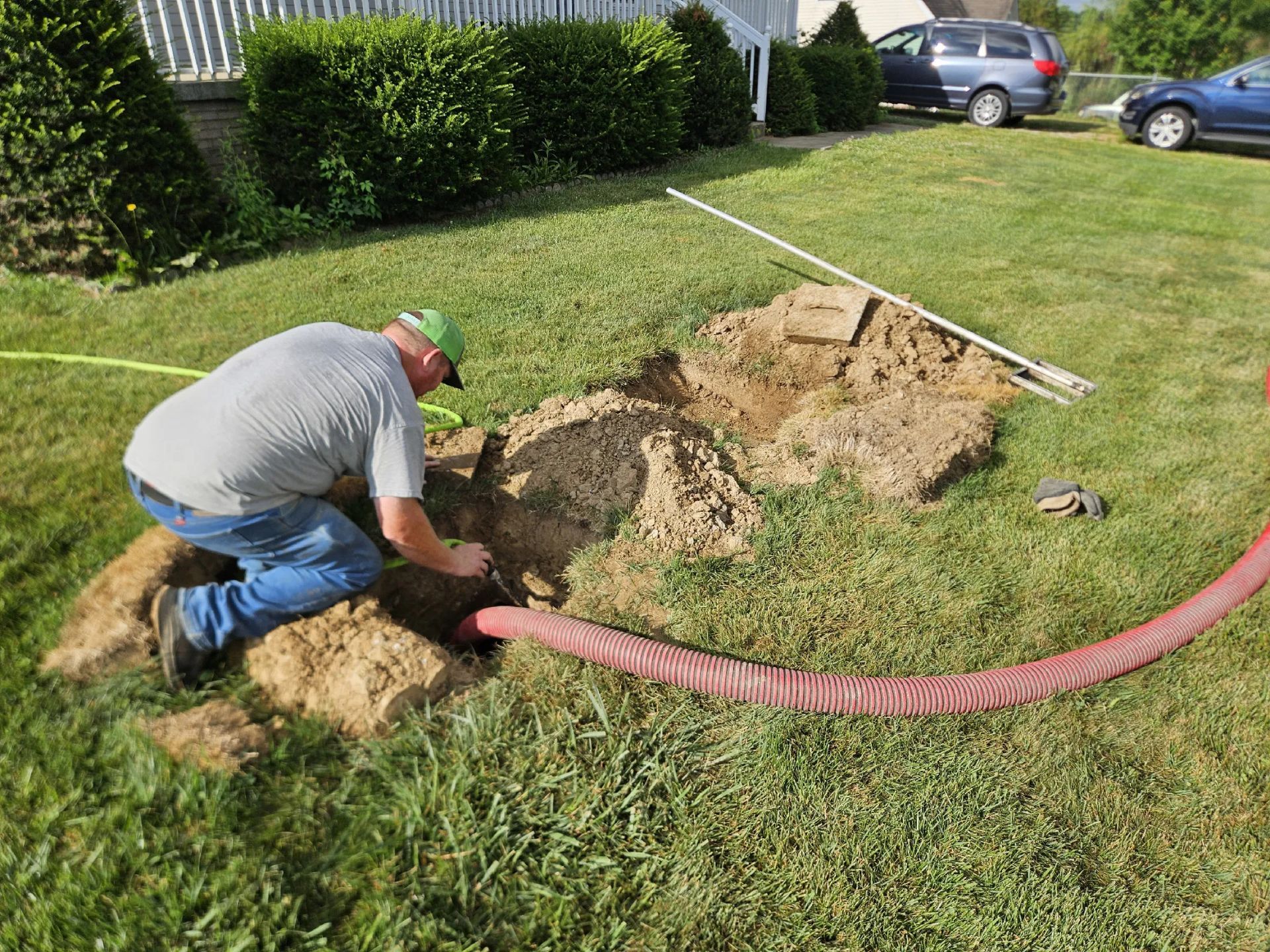 Worker digging trench in a lawn with a pink hose nearby and a mound of dirt beside him