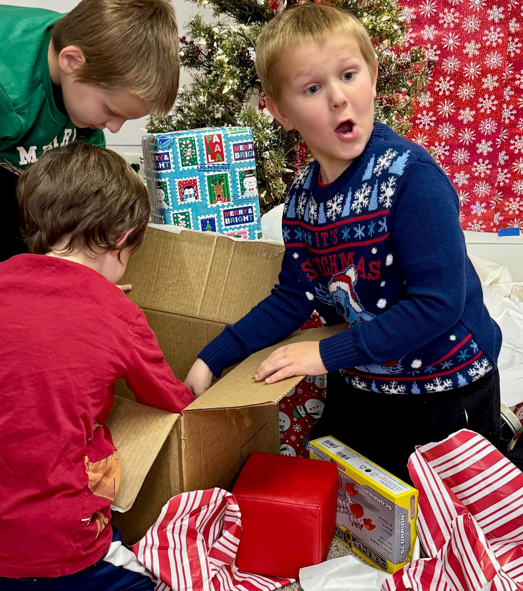 Three young boys are opening christmas presents in front of a christmas tree.