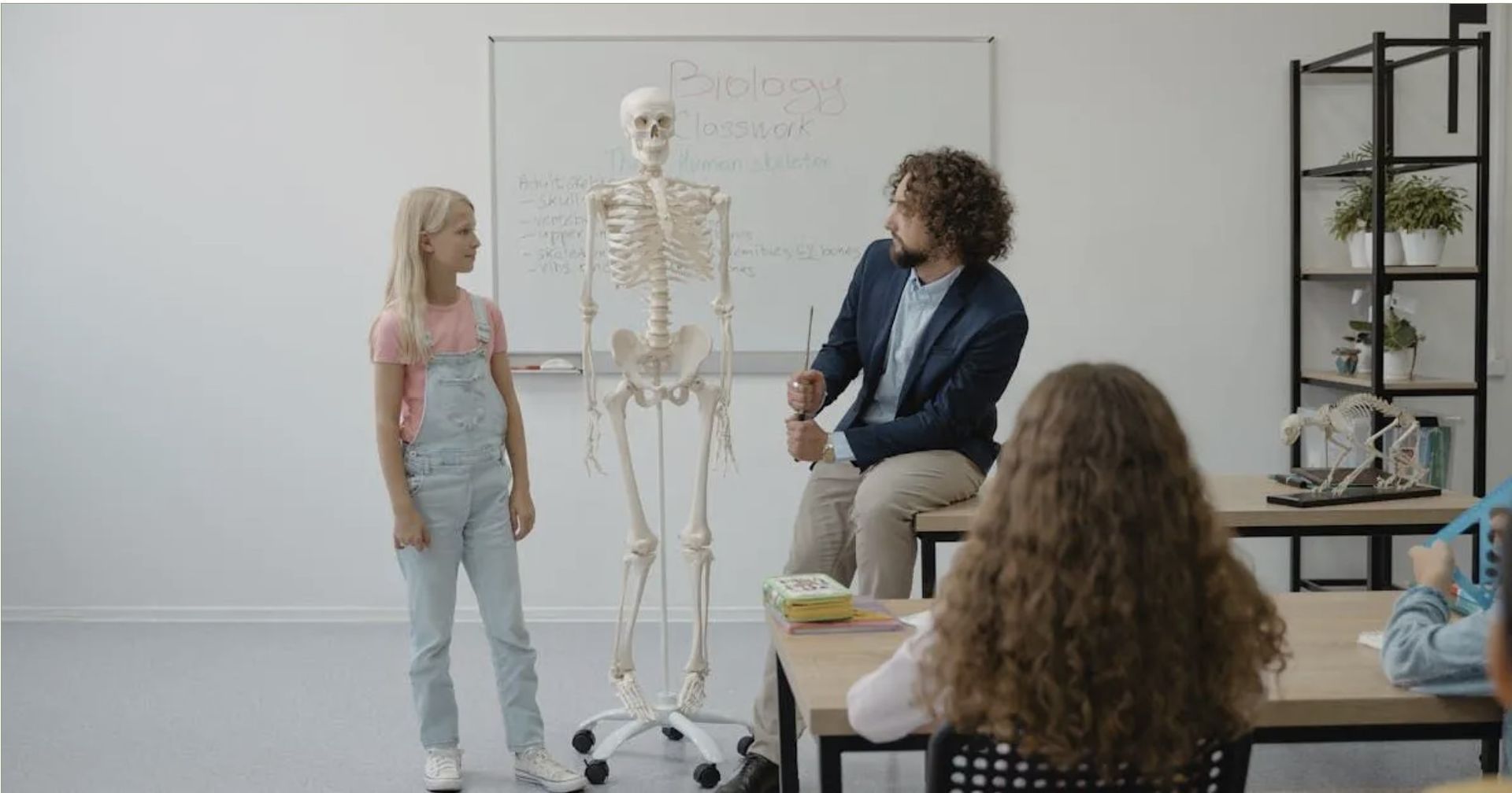 Teacher pointing at skeleton in classroom, girl looking, students at desks.