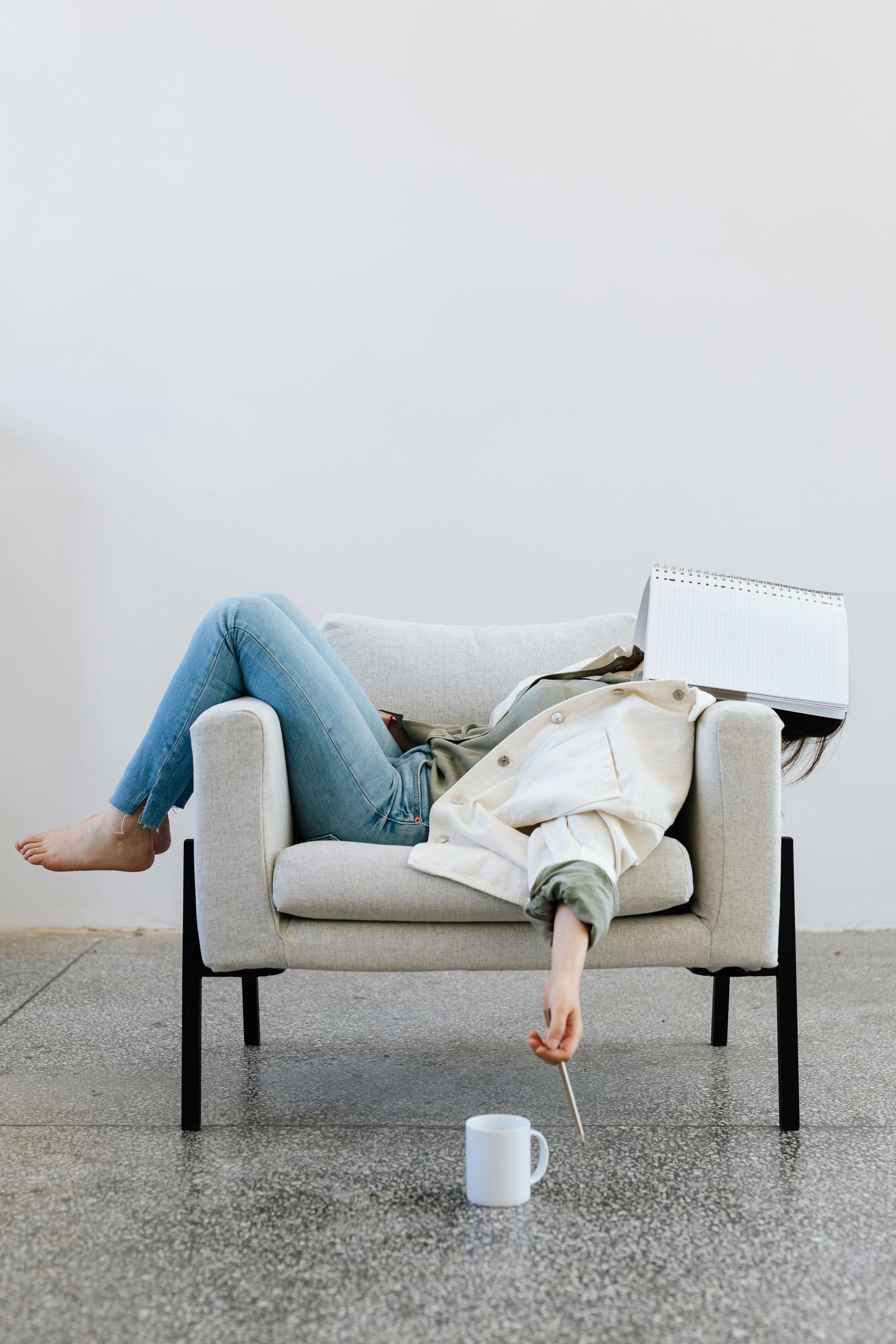 Person slumped in armchair, face obscured by jacket. A hand dangles towards a mug.