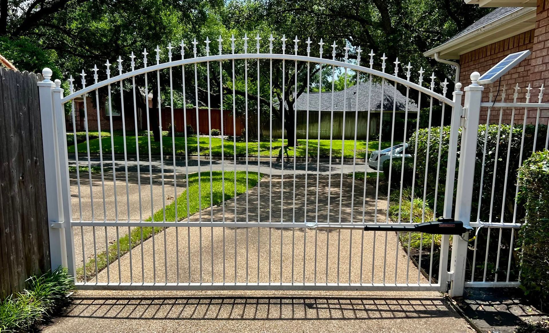White metal driveway gate with arched top, closed in front of a brick house and landscaped yard.