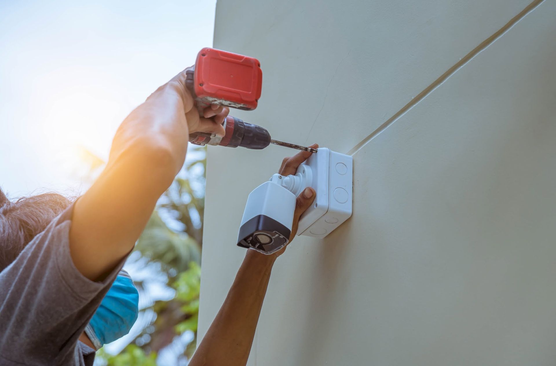 Hands installing a white security camera on a beige wall, holding a red tool nearby.