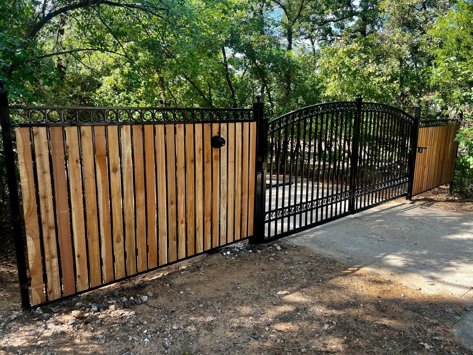 Wooden and black metal driveway gate beside a paved road, with trees in the background.
