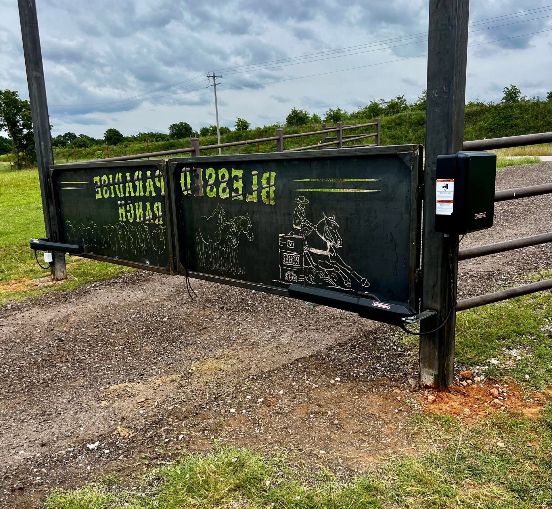 Dark green livestock gate with faded lettering beside a gravel driveway under cloudy sky