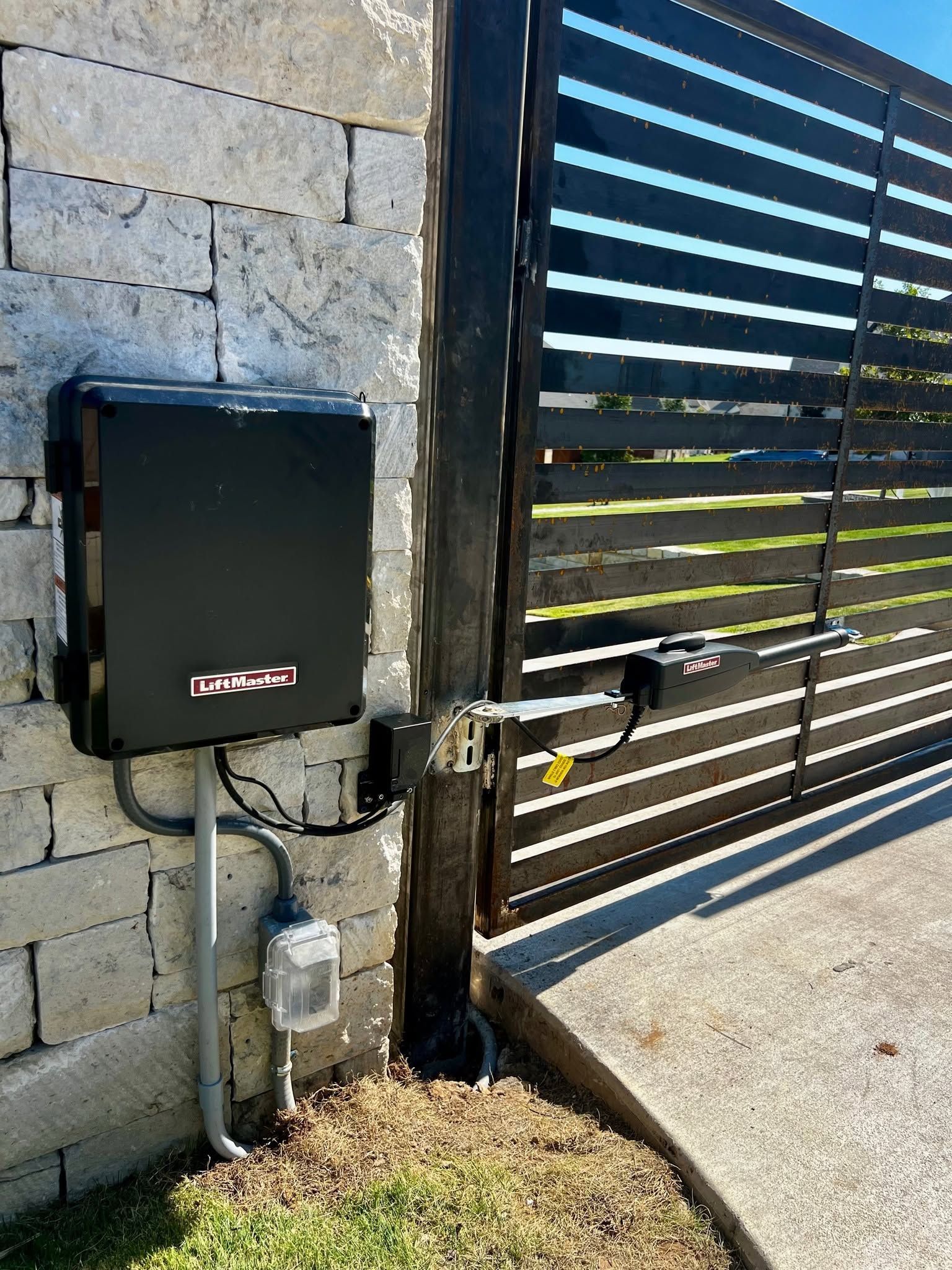 Black utility box mounted on a stone wall beside a slatted metal gate and sidewalk