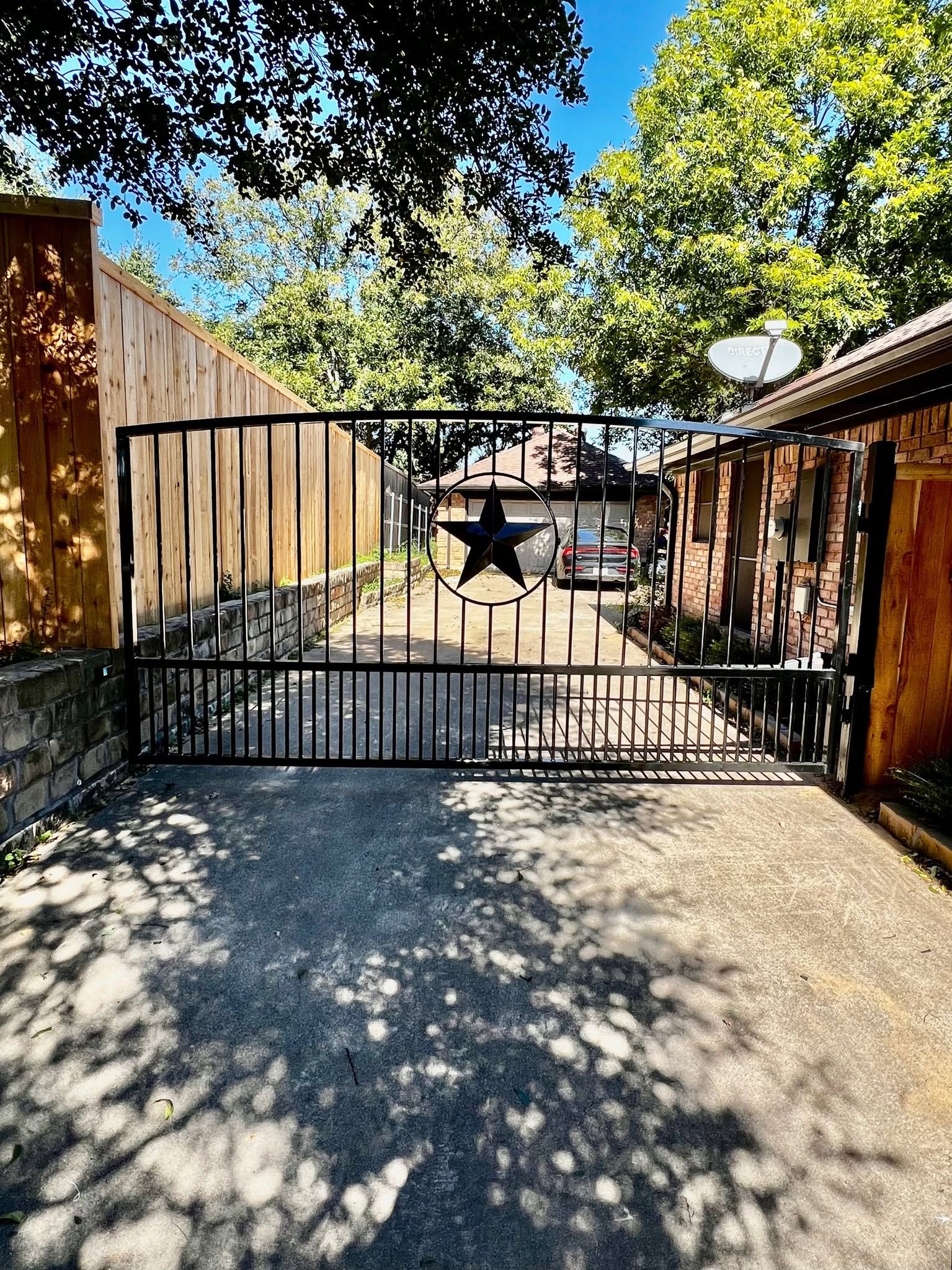 Black metal gate blocking a gravel driveway, with wooden fences and sunlit trees beyond.