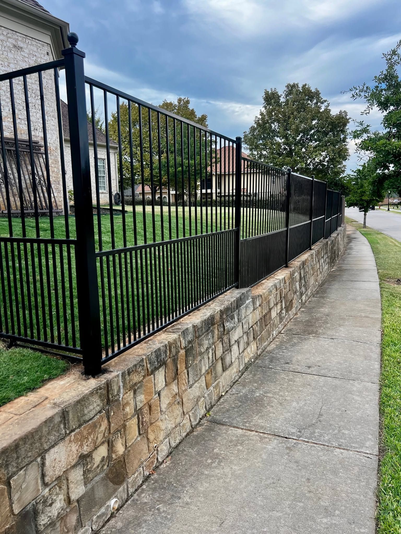 Sidewalk beside a stone wall and black iron fence along a grassy slope on a cloudy day