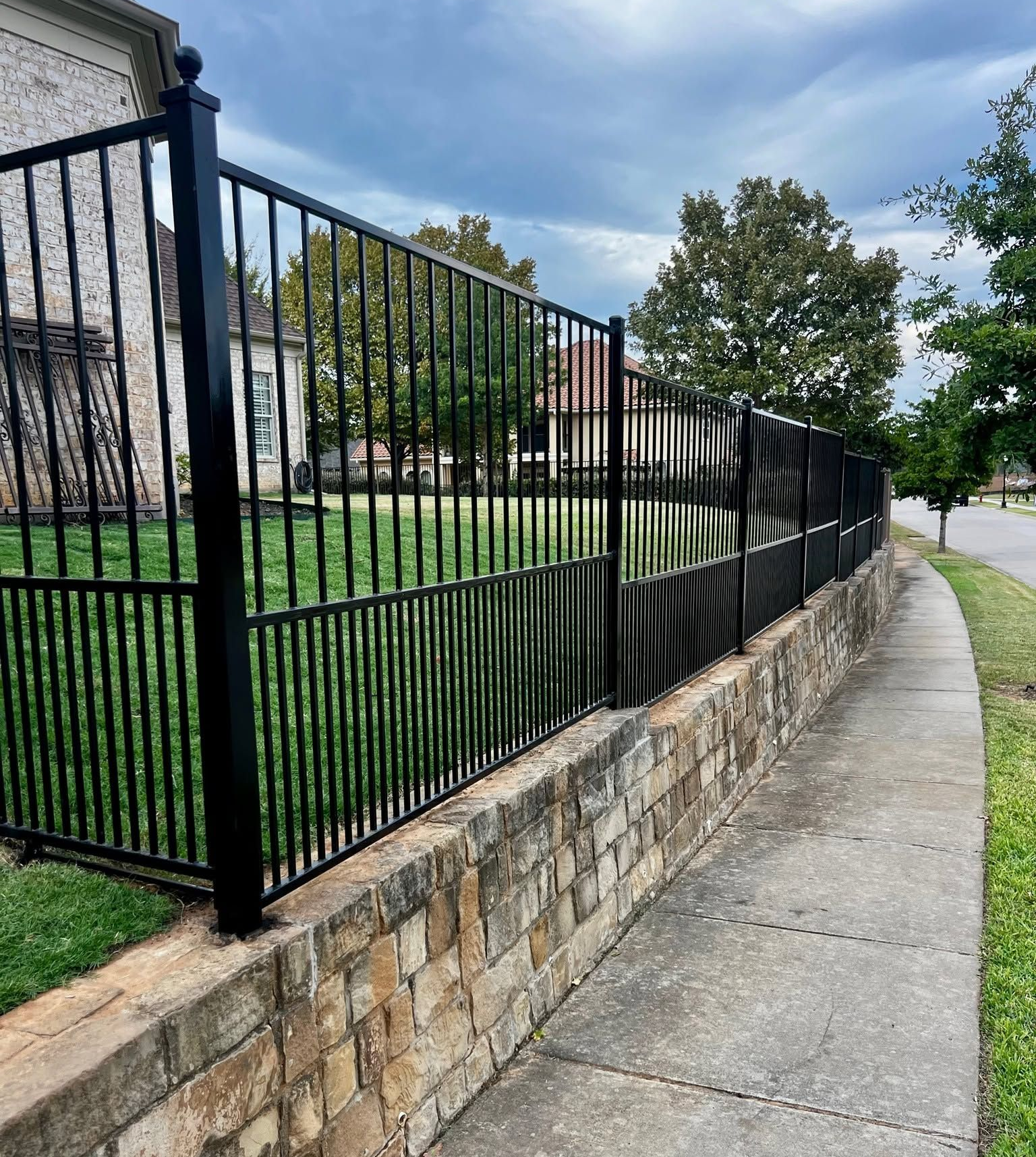 Concrete sidewalk beside a black metal fence and stone wall along a grassy building lawn under cloudy skies