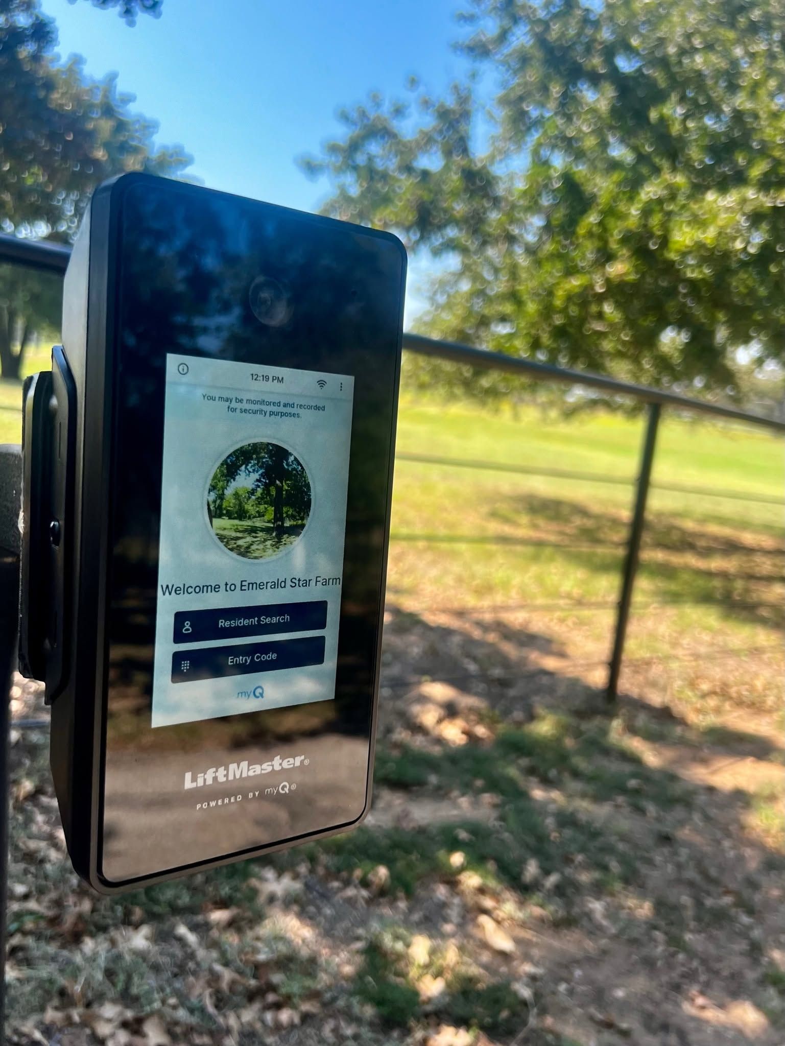Black hand sanitizer dispenser with sign in sunny park by a fence and trees