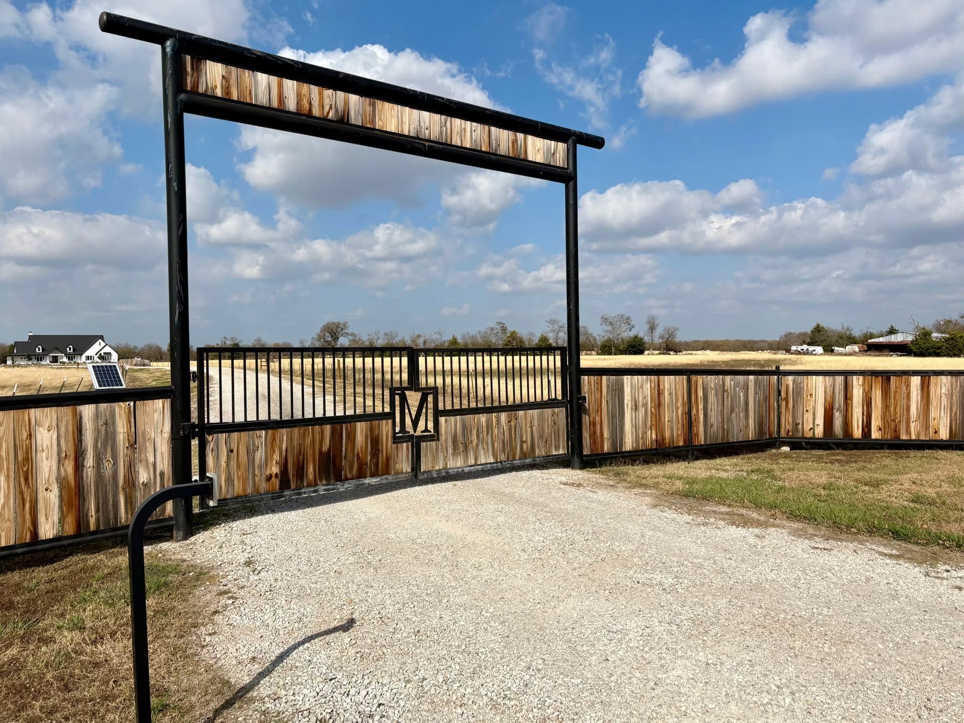 Rustic wooden driveway gate with black metal frame and a latch, beside a gravel path under a blue sky.