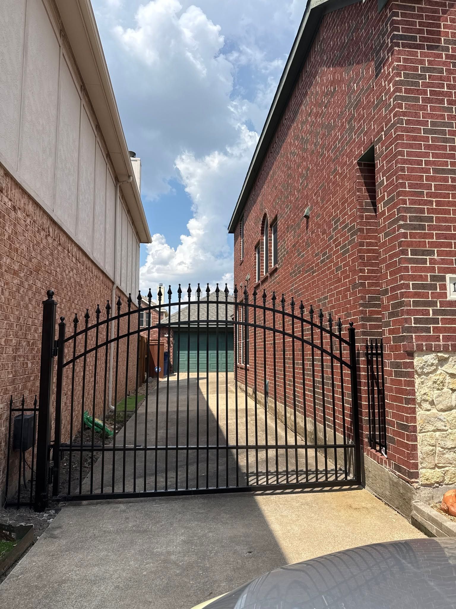Black metal gate between brick buildings, opening to a small yard under a cloudy sky.