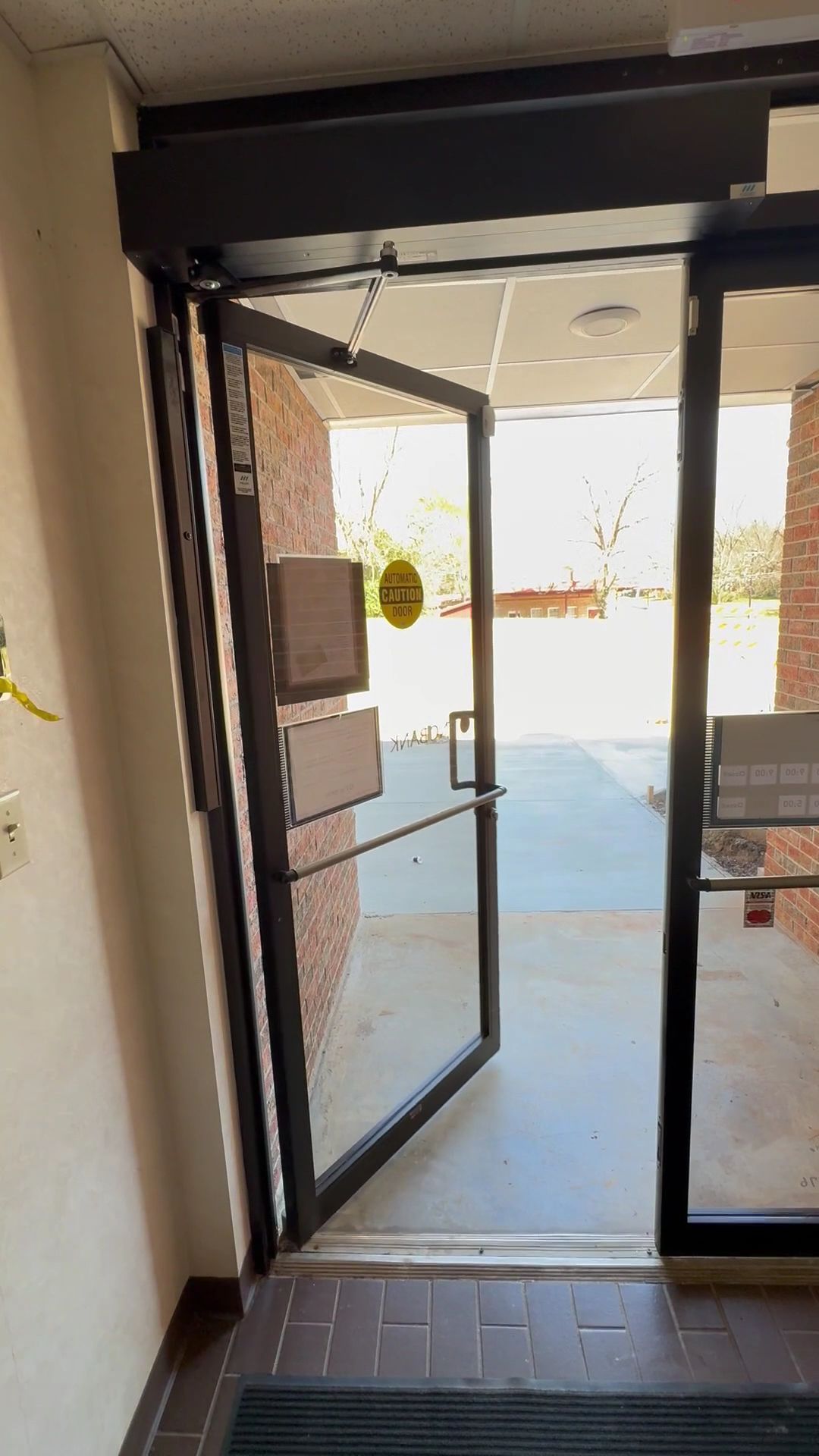 Open glass entrance door leading to a bright outdoor walkway from an indoor lobby.